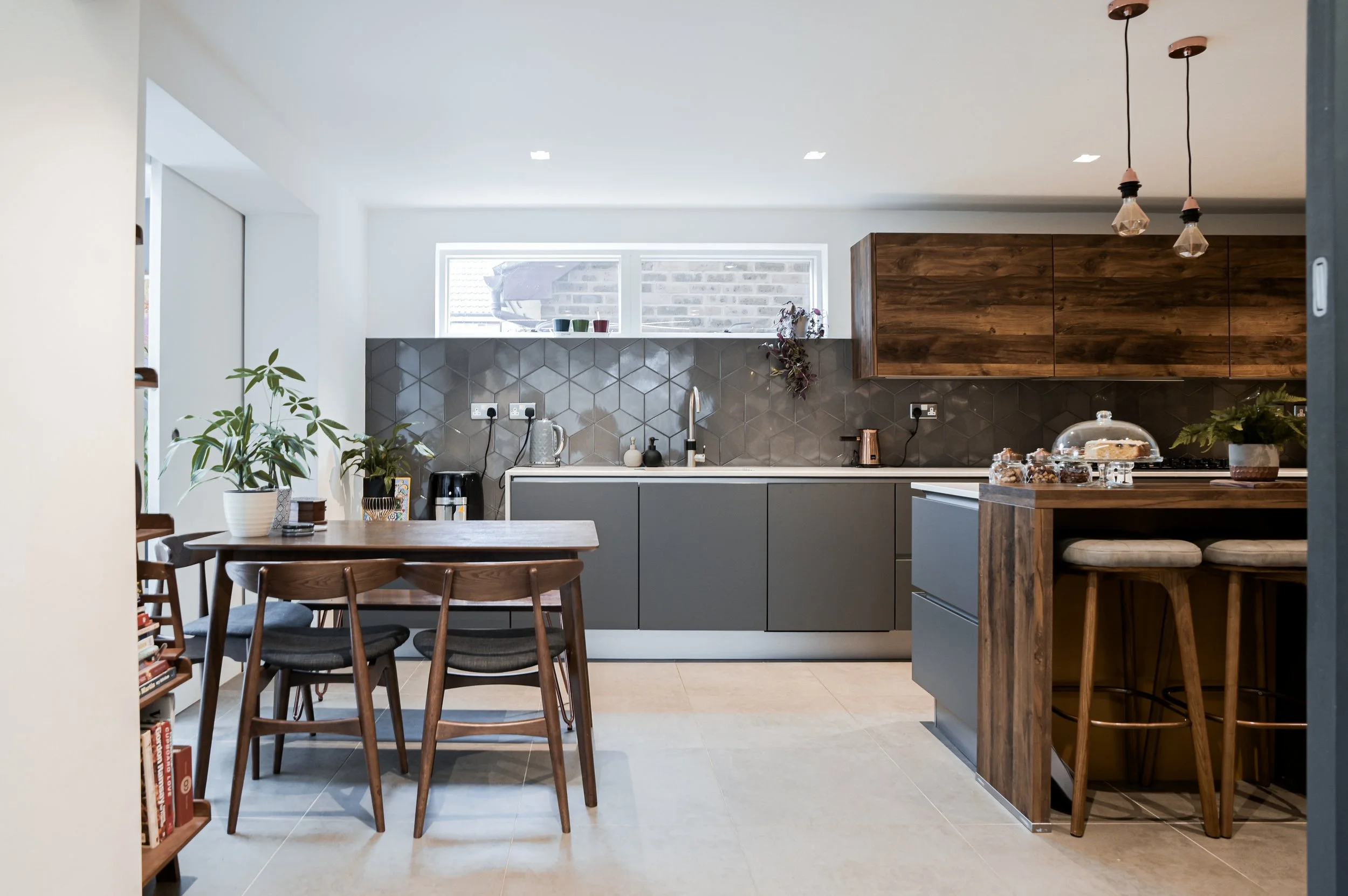 Modern kitchen with gray cabinets, wooden accents, and geometric tile backsplash. There is a small dining table with chairs, potted plants, and decorative items on the counter, with pendant lights hanging from the ceiling.