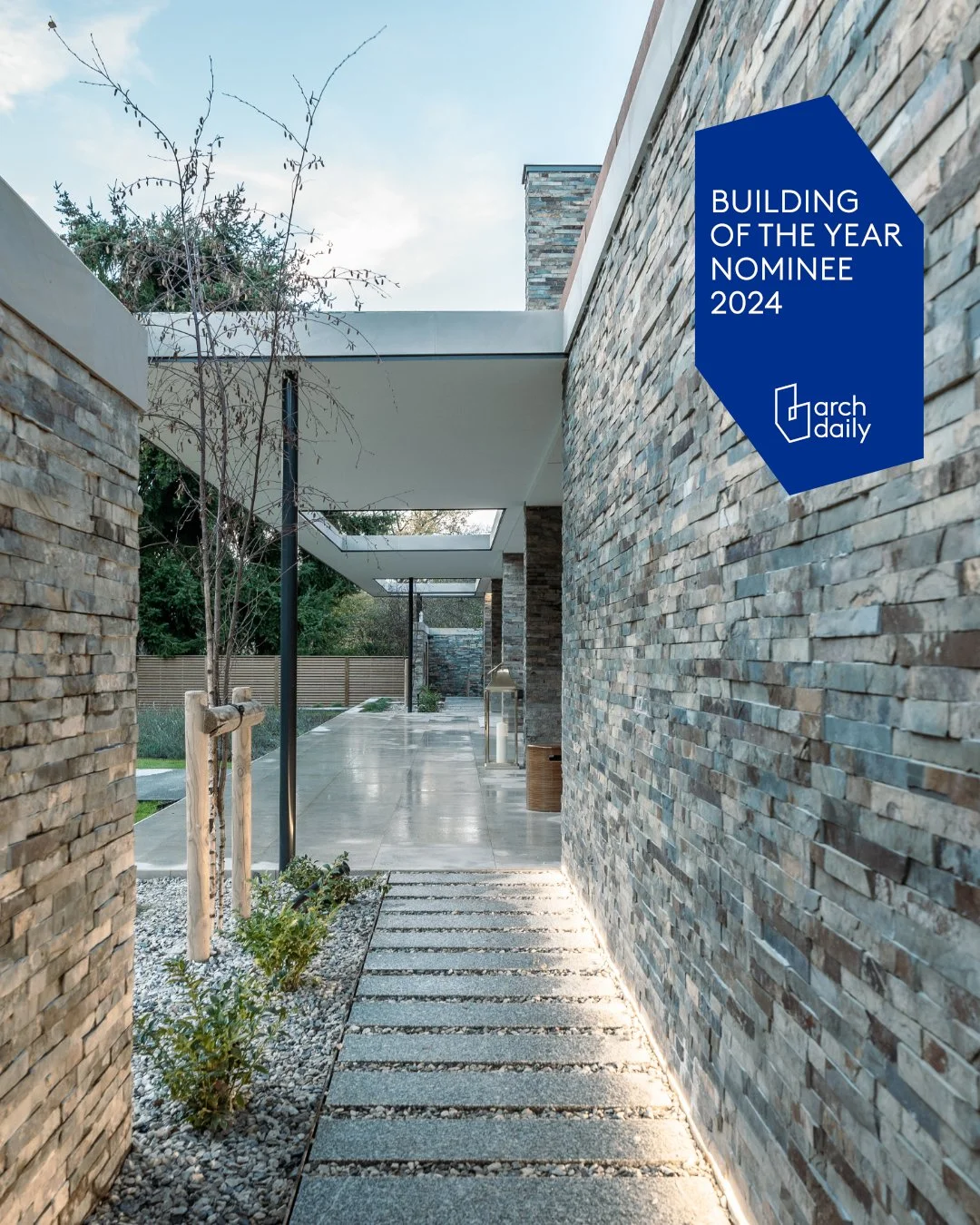 Modern outdoor corridor with stone walls, a pathway made of rectangular tiles and gravel, under a white canopy, with trees and greenery in the background, and a blue sign indicating it is a 2024 building of the year nominee by arch daily.