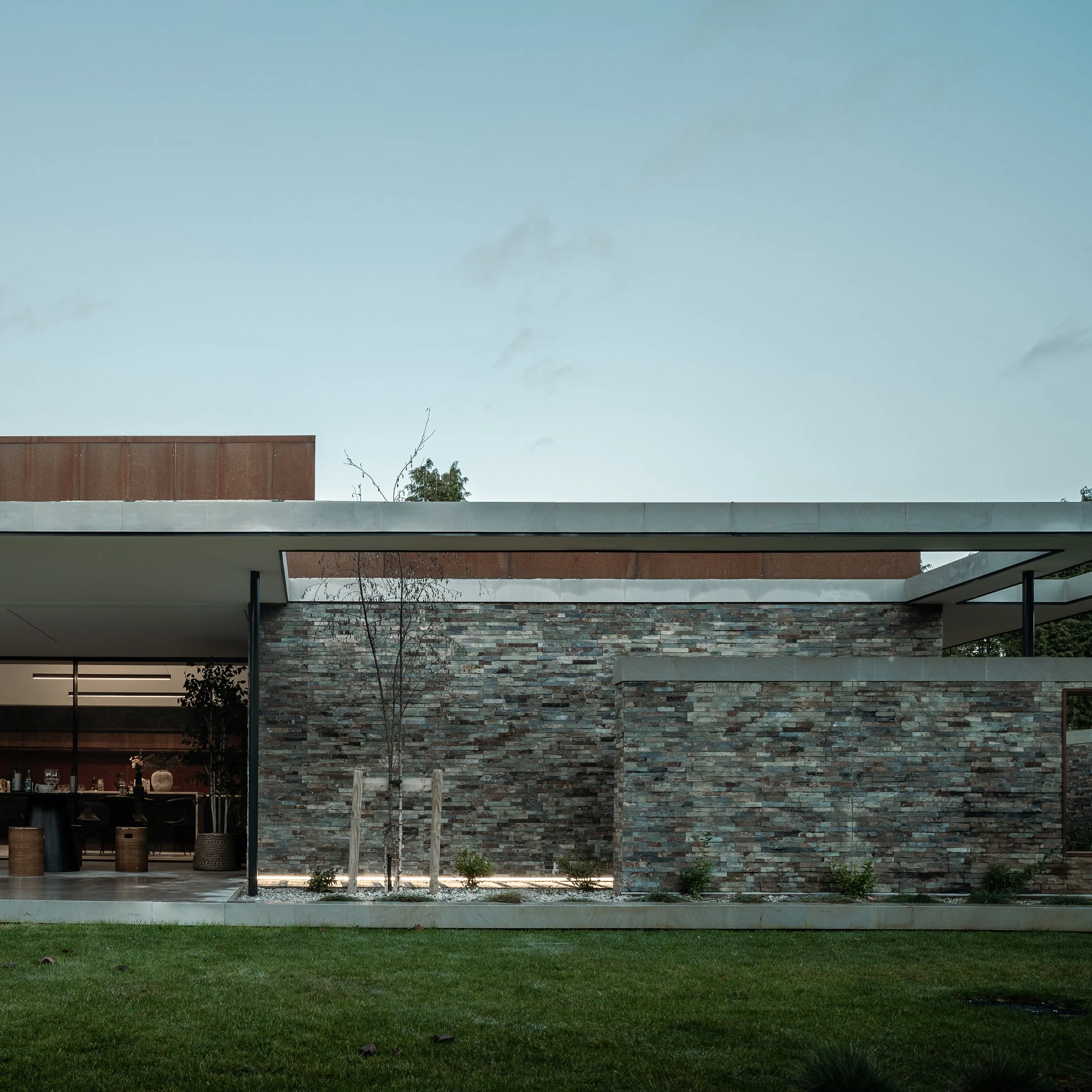 A modern house exterior with a brick and stone facade, large windows, and a patio area with outdoor furniture, surrounded by a neatly maintained lawn and young trees.