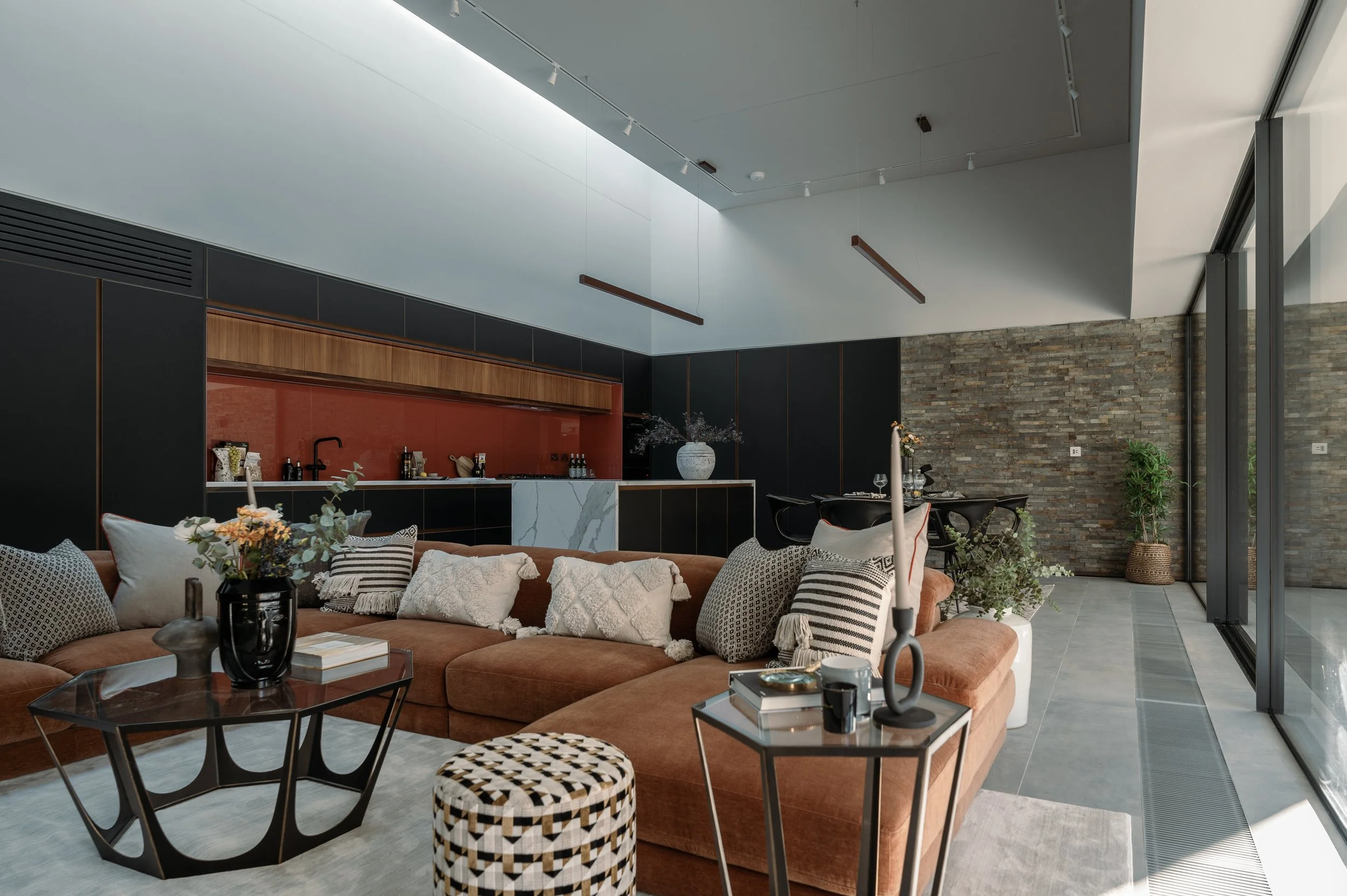 A modern living room with a large L-shaped sectional sofa with cushions and throws, a glass coffee table, and a small round cushioned stool with a black and cream pattern. The background features a black kitchen area with wooden accents, a red backsp
