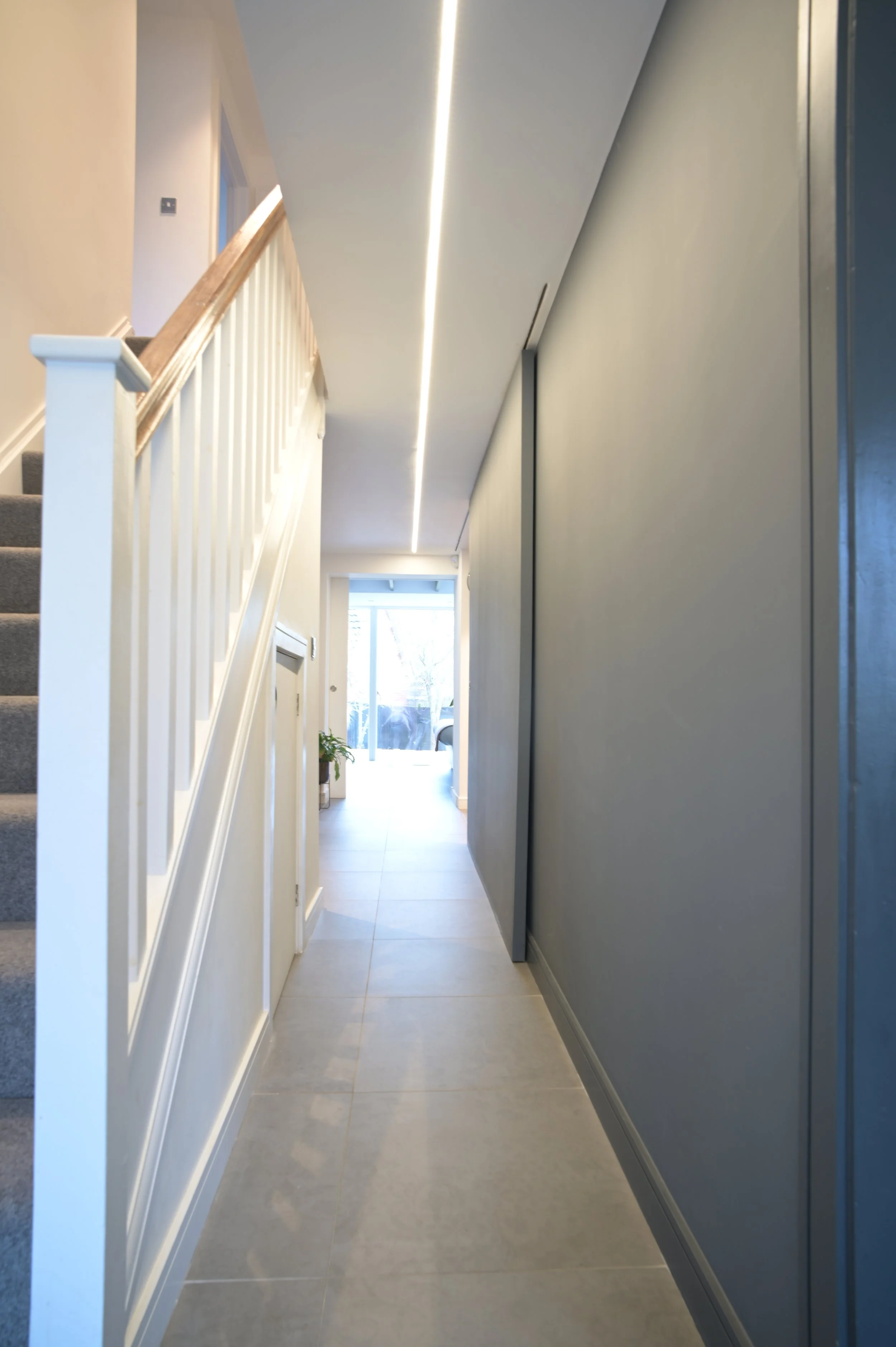 A modern hallway with a staircase on the left, a tall gray cabinet or wall on the right, and a large window at the end providing natural light.