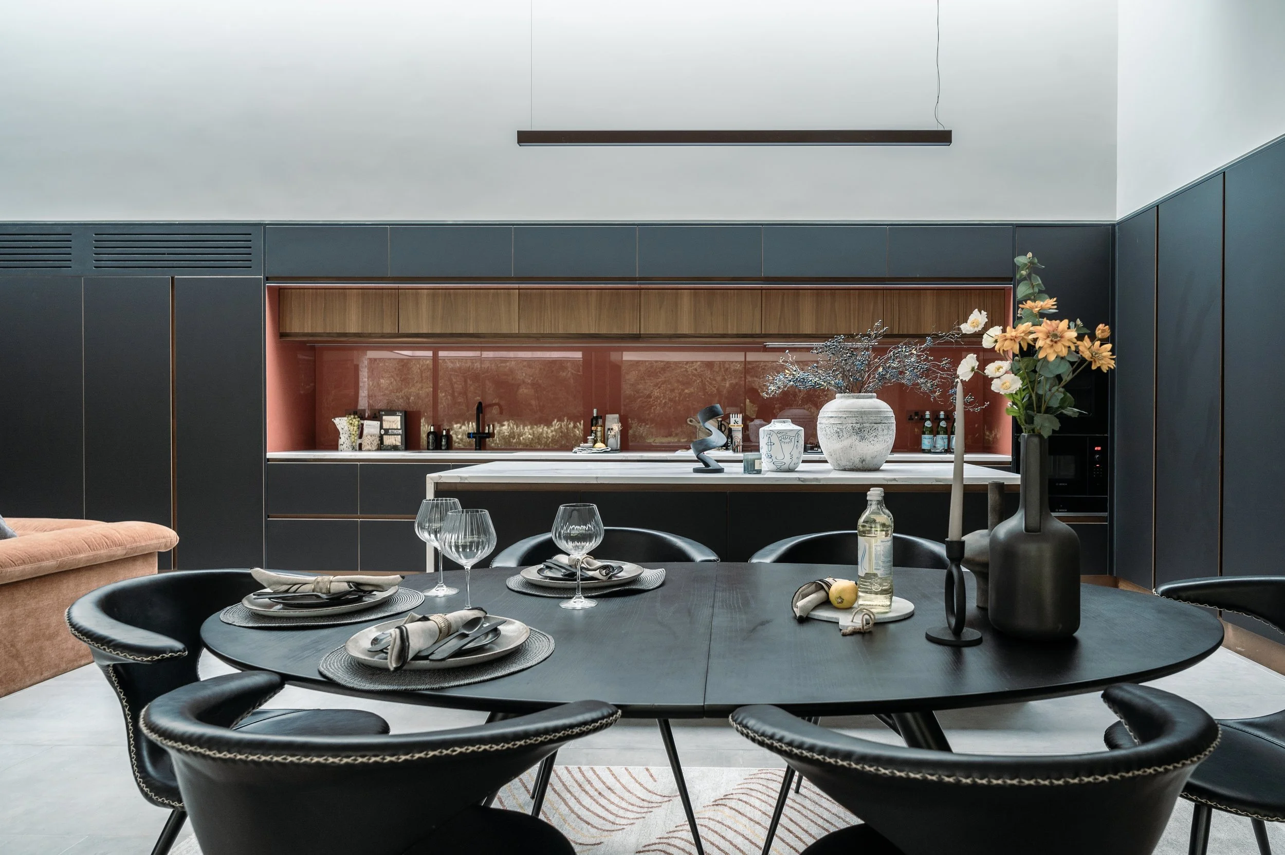 Modern dining room with a black round table set with glasses, napkins, and plates, black chairs, floral decorations, and a kitchen with dark cabinetry, wooden accents, and a pink backsplash.
