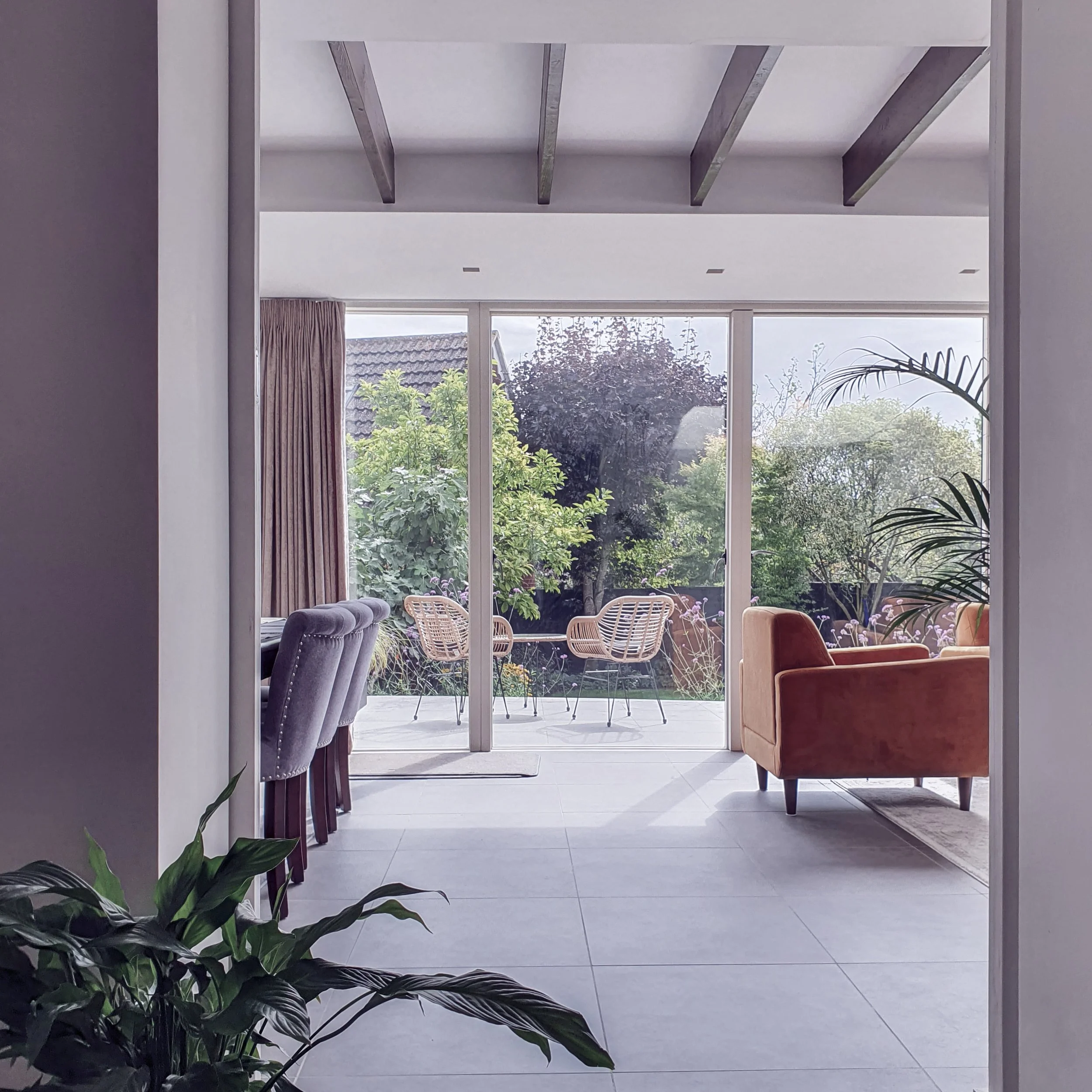 View through a glass door to an outdoor patio with two wooden chairs, lush green plants, and trees under a partly cloudy sky.