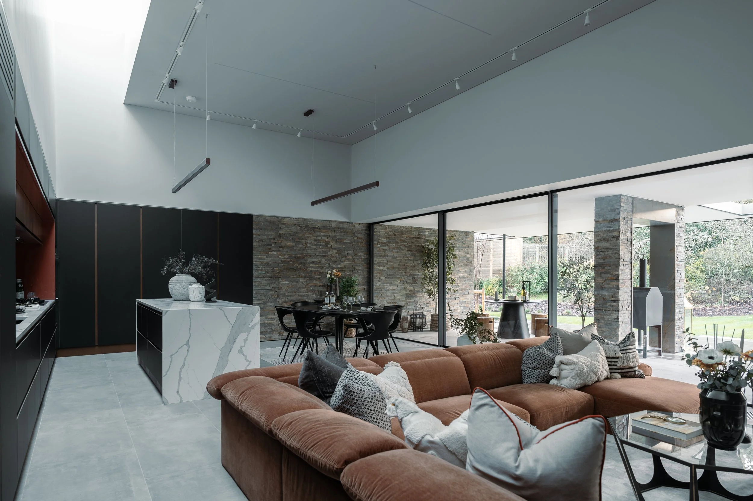Modern open-concept living and dining area with large floor-to-ceiling windows, a brown sofa with patterned pillows, a black dining table with chairs, a stone accent wall, and a marble kitchen island