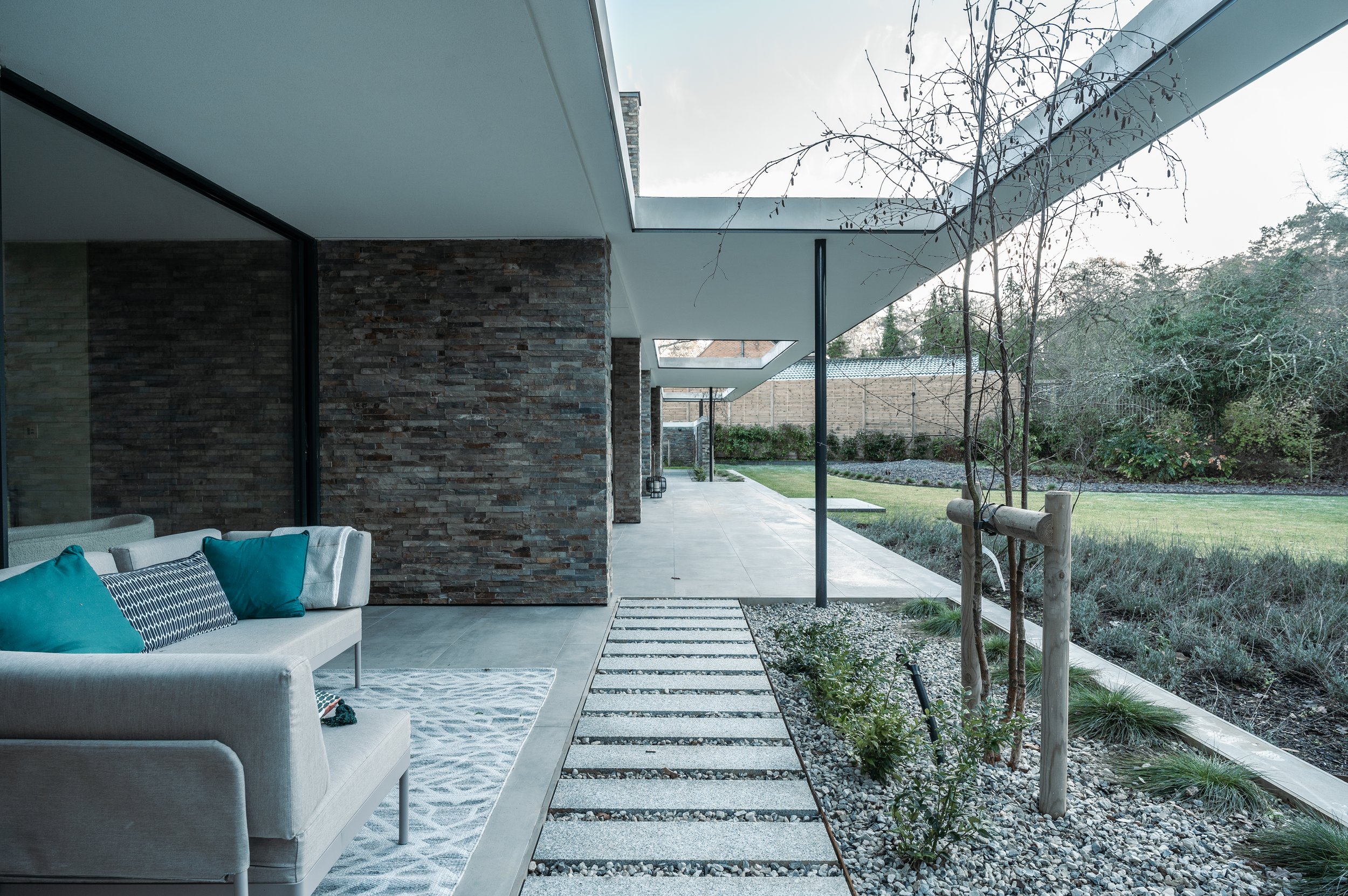 Modern house exterior with a covered patio, stone wall, and a view of a backyard with trees and a lawn.