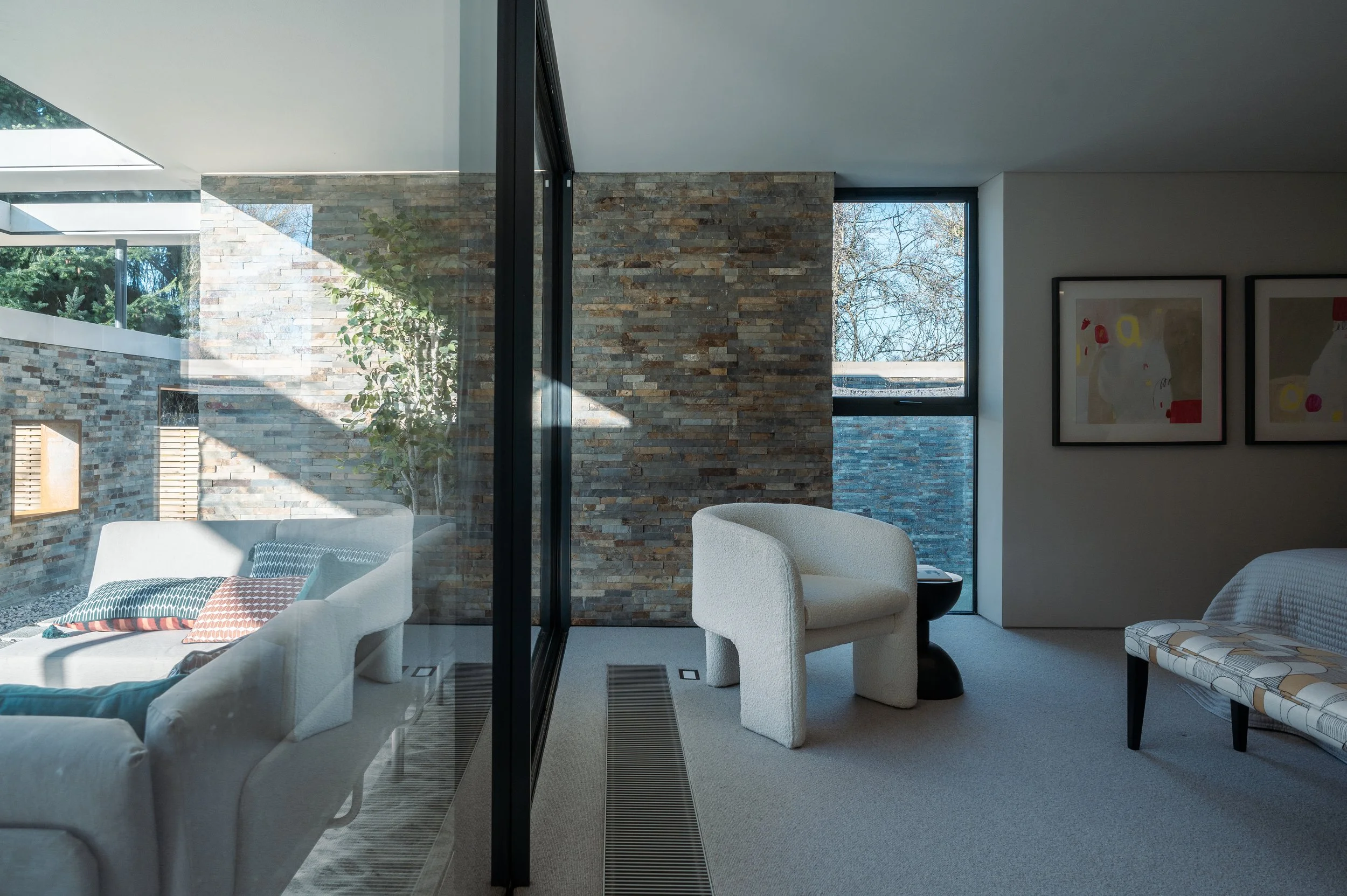 Modern living room with white sofa and colorful pillows, large windows, and a stone wall visible through a glass partition.