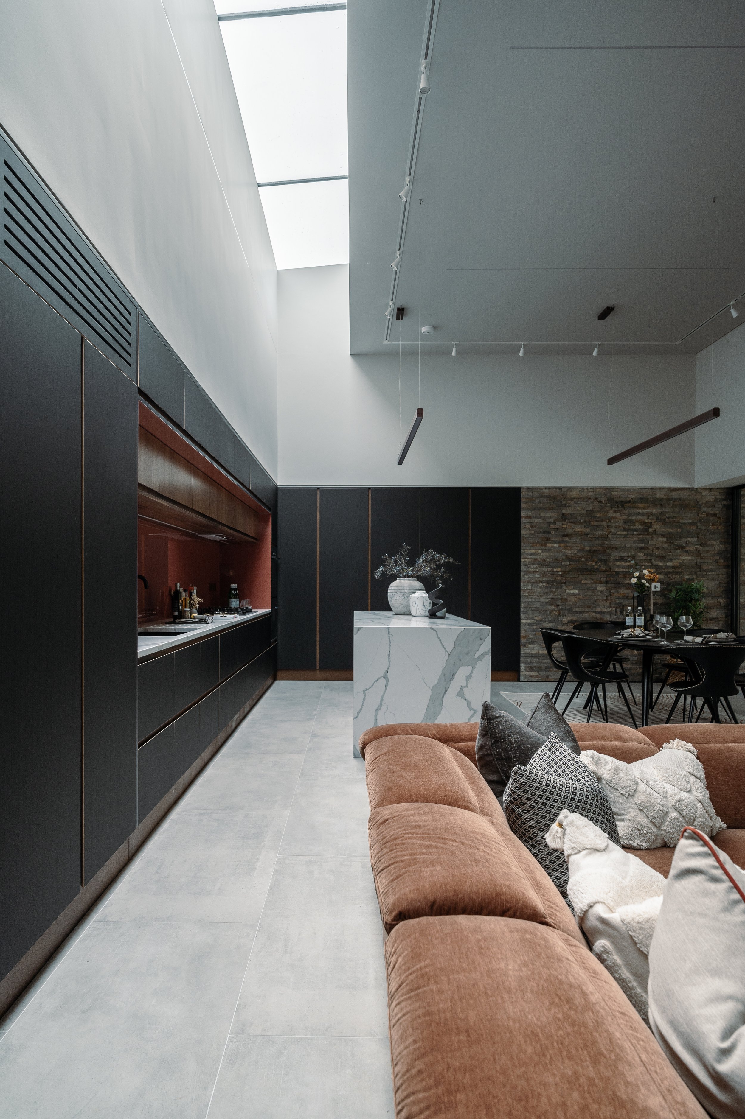 Modern living room with a brown velvet sofa, decorative pillows, a white marble table with vases, black dining chairs with wine glasses, and a brick accent wall.