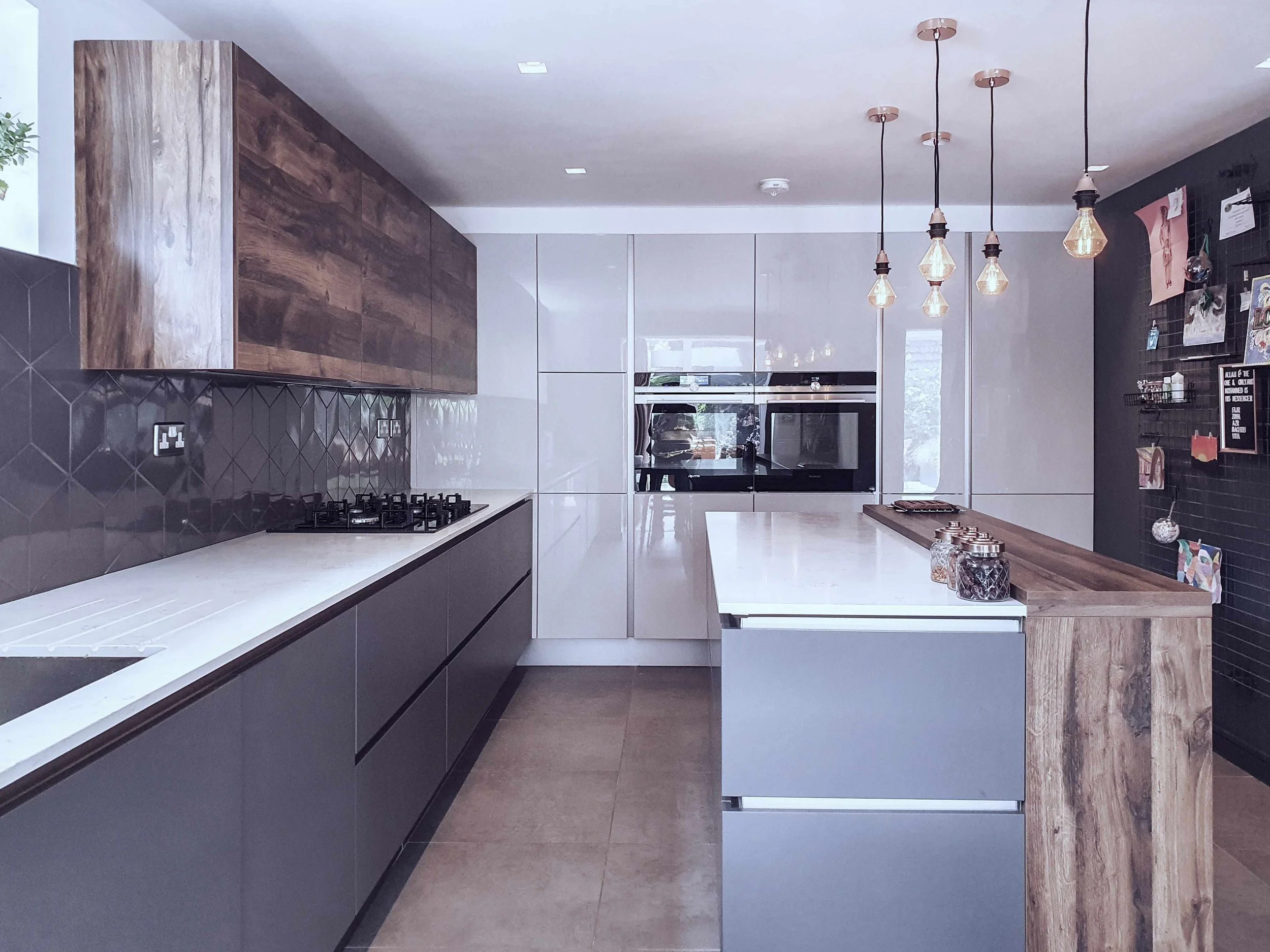 Modern kitchen with white countertops, wooden accents, and pendant lighting, featuring a black tile wall with notes and decorations.