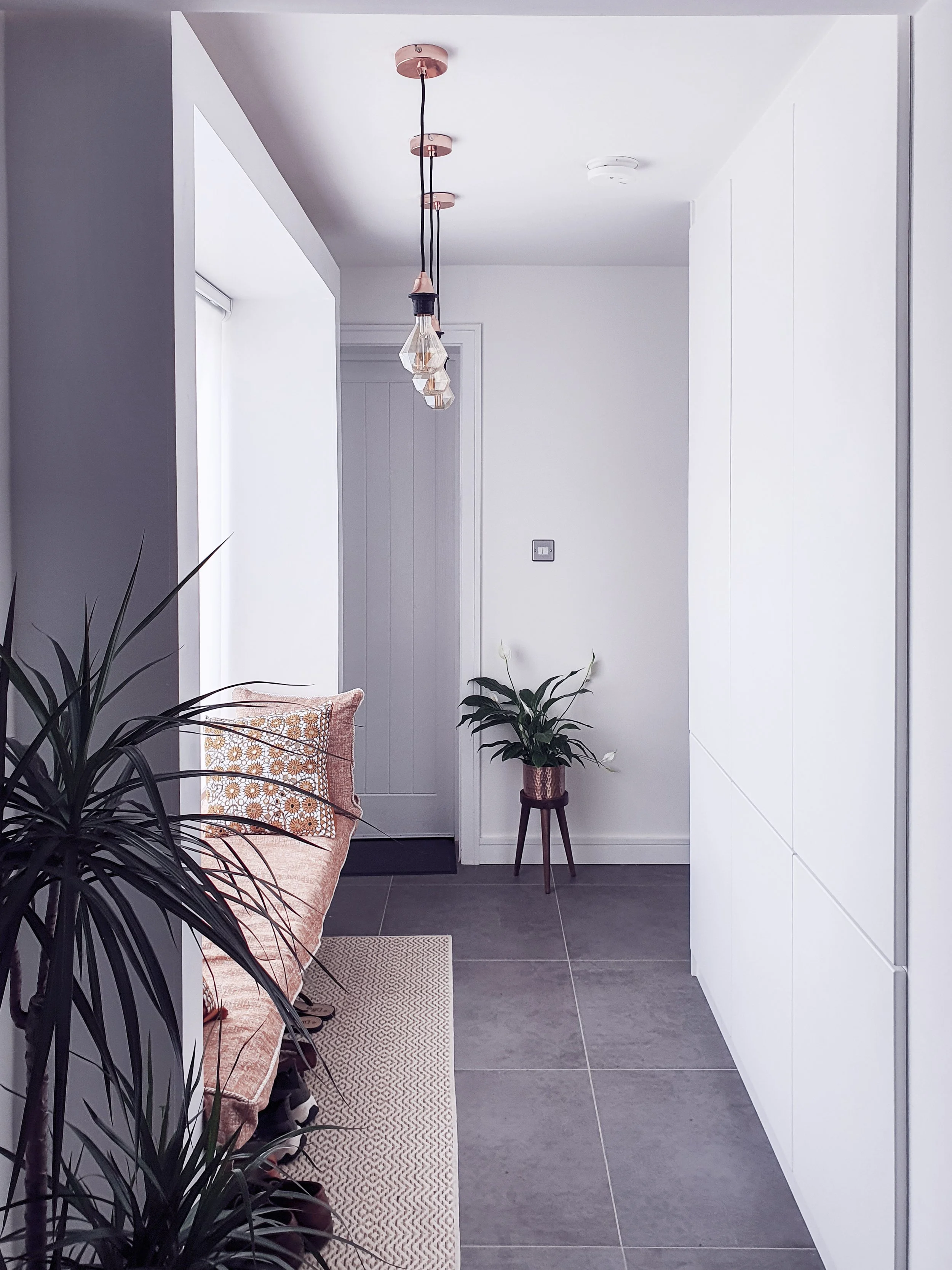 A modern, minimalist entryway with white walls, gray tiled floor, a peach-colored sofa with patterned cushions, and potted plants. A hanging light fixture with three bulbs is visible on the ceiling.