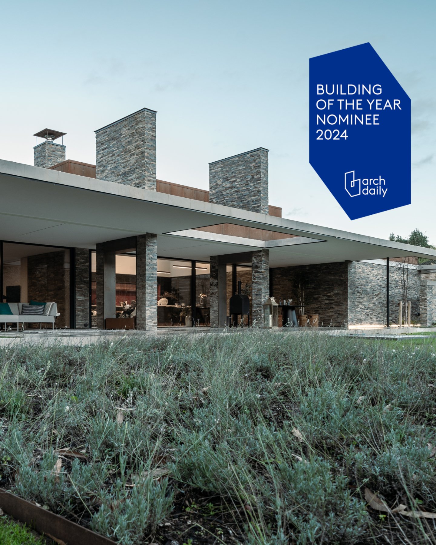 Modern house with stone chimney and large glass windows, surrounded by grass and landscaping, with a blue banner indicating it is a 2024 building of the year nominee.