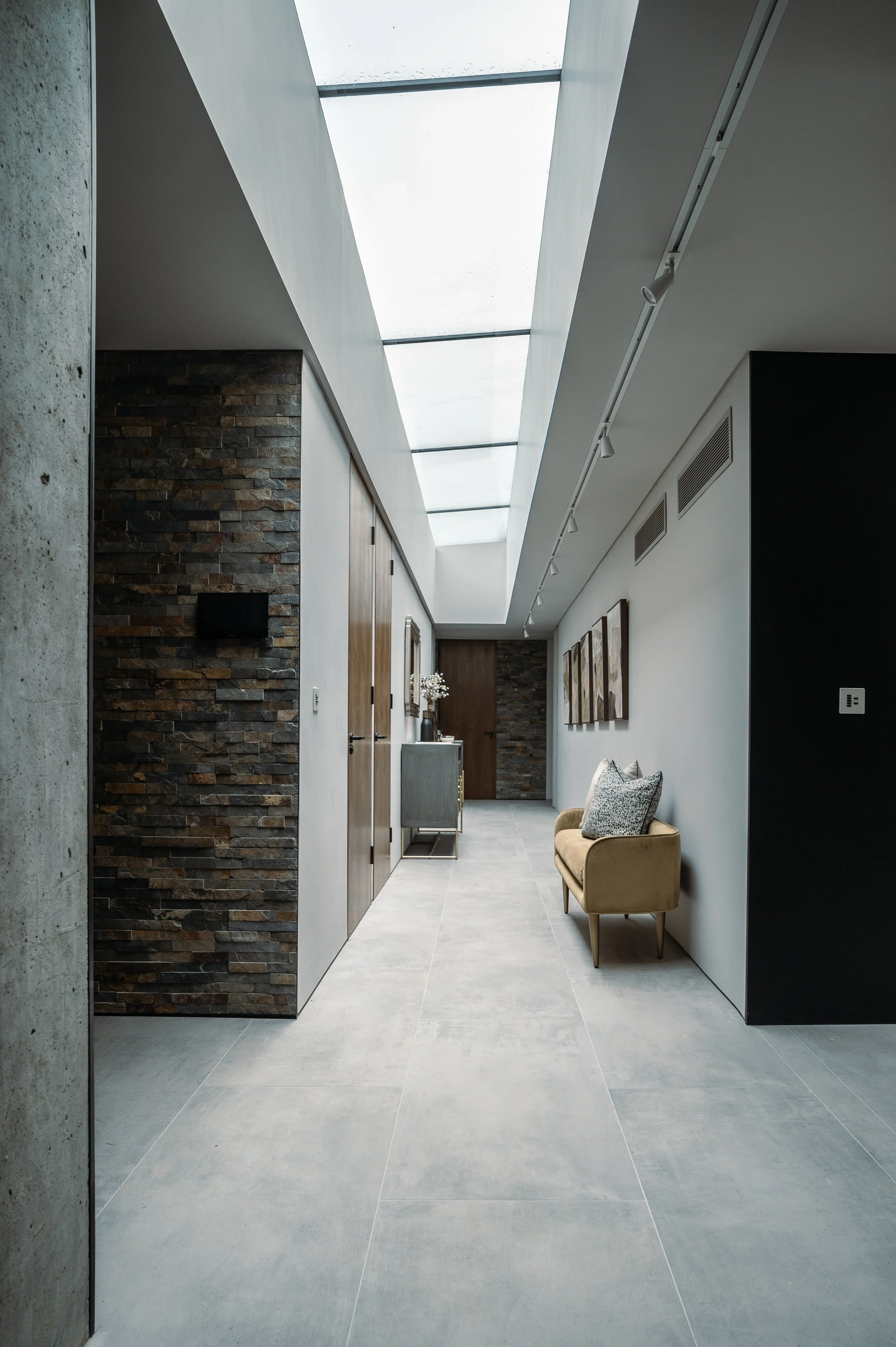 Modern interior hallway with large skylight, stone and white walls, a beige bench with cushions, wall art, and a small table with a vase of flowers.