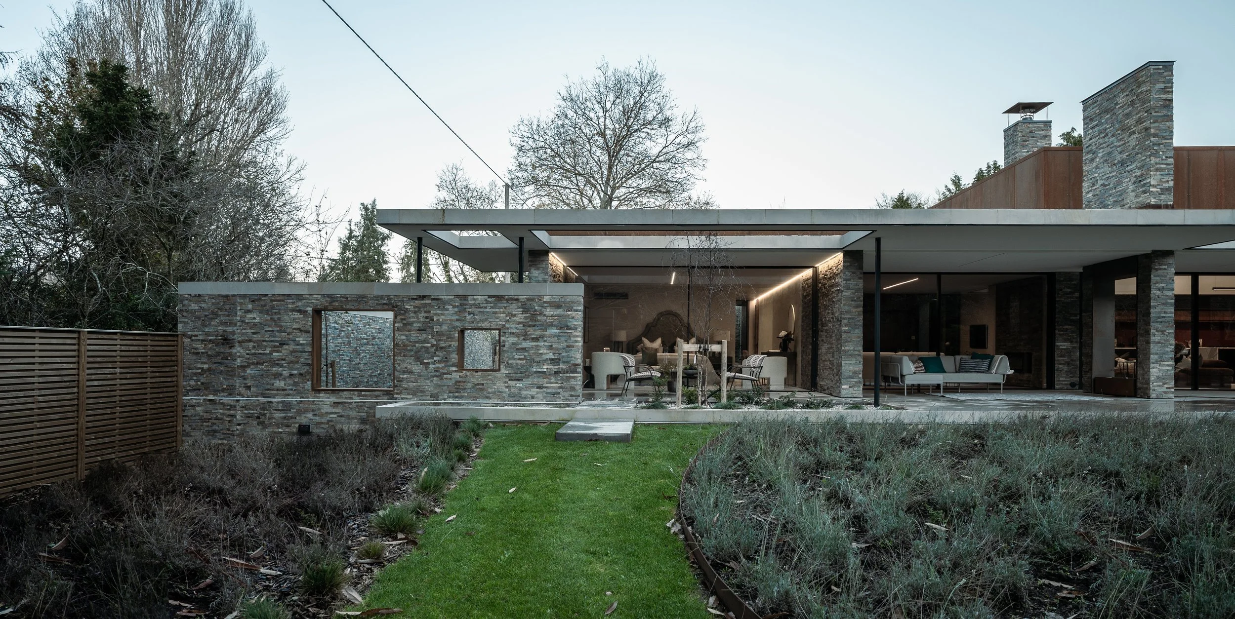 Modern house with large covered patio, glass sliding doors, and stone exterior walls, surrounded by trees and a garden.