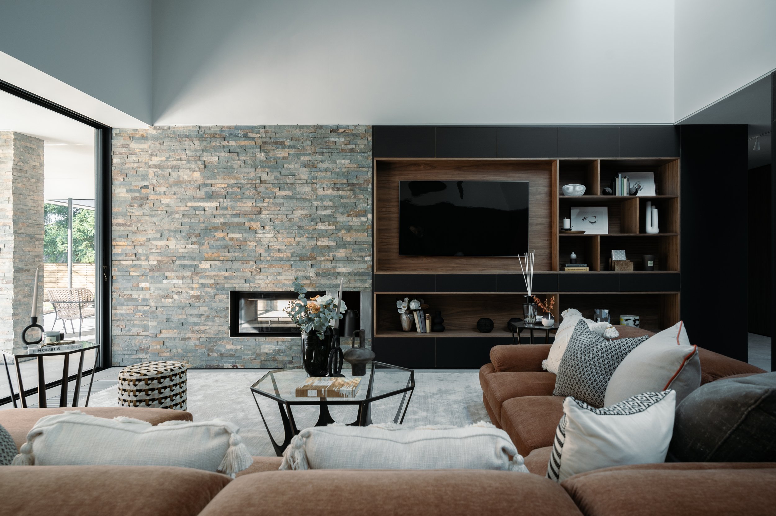 Modern living room with a large stone accent wall, a built-in wooden and black TV unit, a cozy brown sofa, and a glass coffee table with decorative items. There's a large sliding door on the left and a mix of minimalistic decor and books.