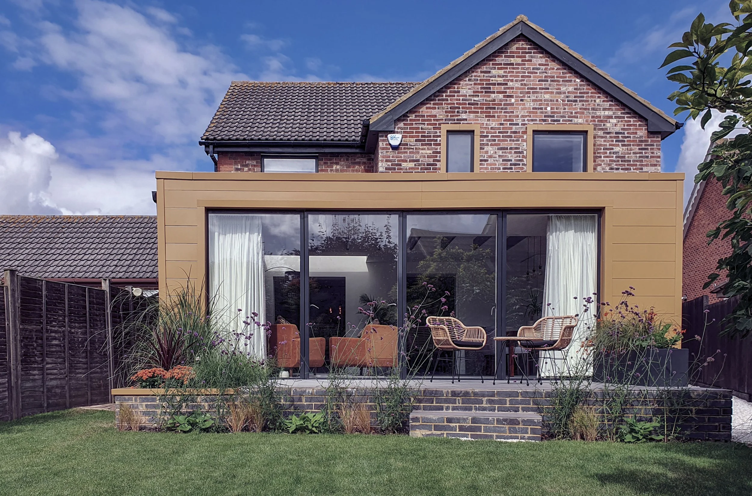 A modern house with a brick upper floor and a beige extension with large glass sliding doors, outdoor furniture, plants, and a green lawn under a partly cloudy sky.