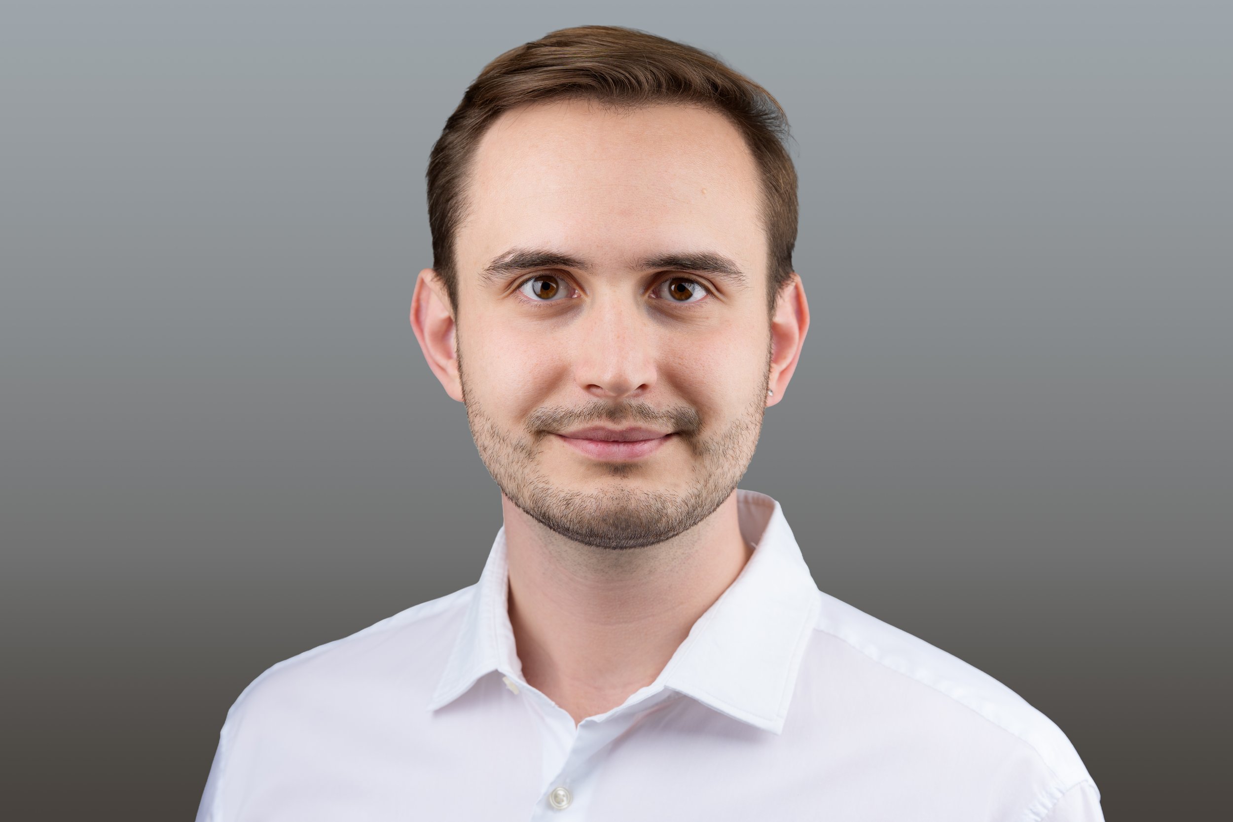 Porträt eines jungen Mannes mit braunen Haaren, braunen Augen, leichtem Bart und einem weißen Hemd vor einem grauen Hintergrund.