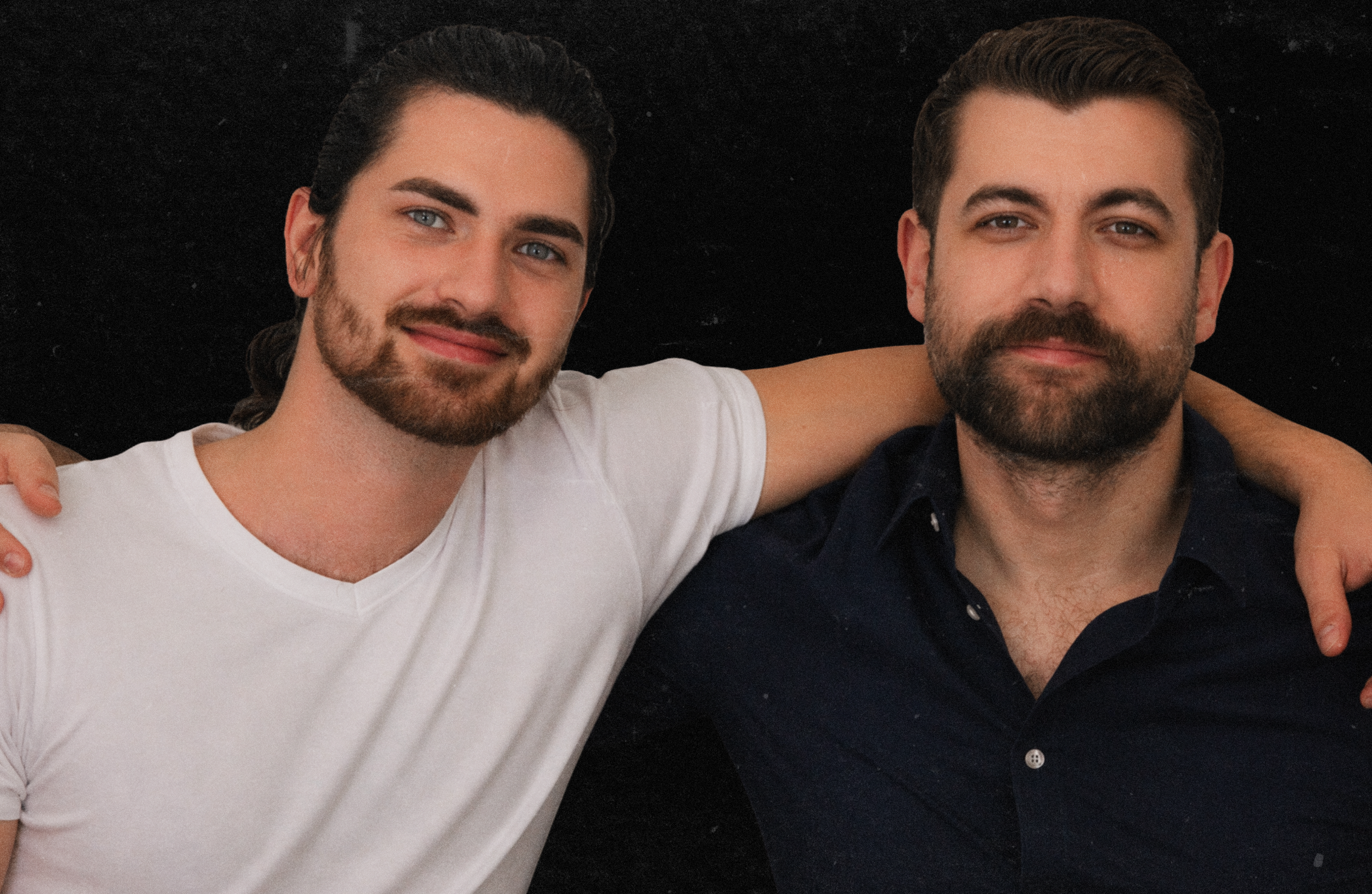 Two young men with beards and short hair, one wearing a white shirt and the other a dark shirt, are smiling and have their arms around each other's shoulders against a dark background.