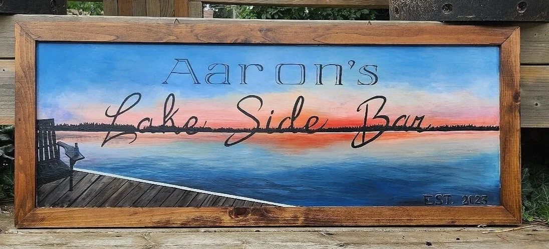 Painted sign for Aaron's Lake Side Bar with a sunset over a lake, a wooden deck, and a black bench, framed in wood, on a wooden surface outdoors.