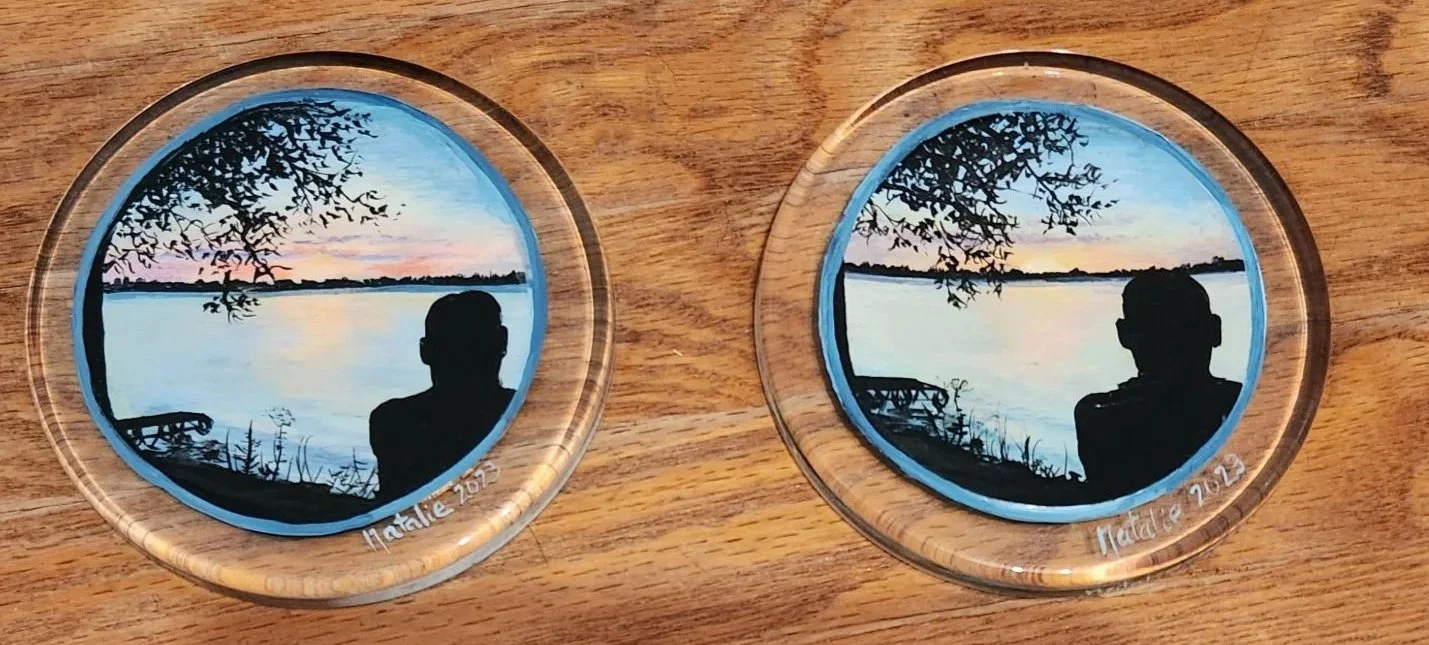 Two round glass coasters with scenic sunset landscapes, silhouettes of a person and tree branches at the lake, signed 'Natile 2022', placed on a wooden surface.