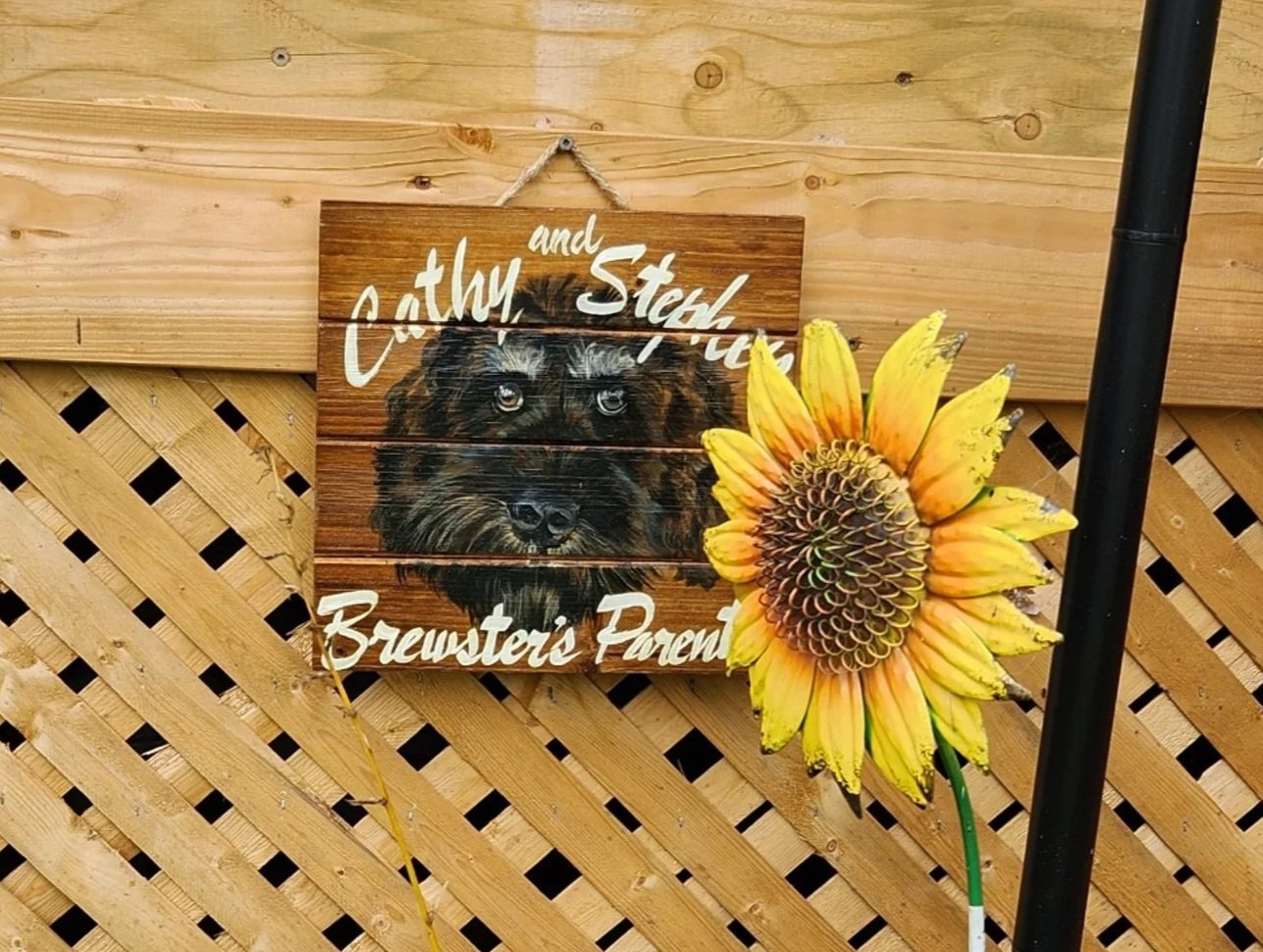 Wooden porch railing with a sign that says 'The Hanna's Home Away From Home', a picture of a dog with the text 'Cathy and Steve Brewster's Pared', a sunflower, and potted plants, with green leafy plants in the foreground.