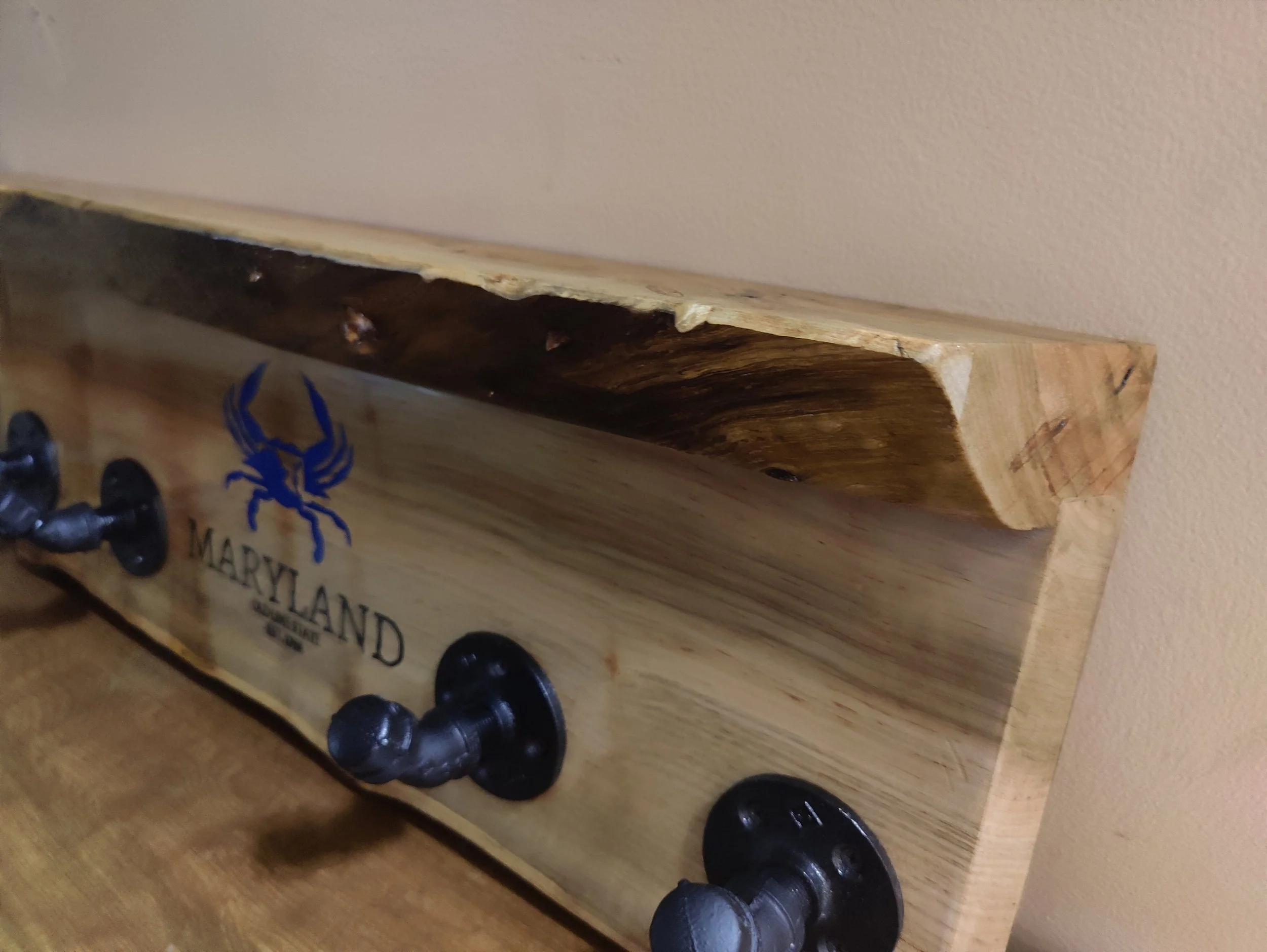 Wooden wall organizer with three black hooks, a blue Maryland crab logo, and the text 'MARYLAND' on the front, mounted on a wall.