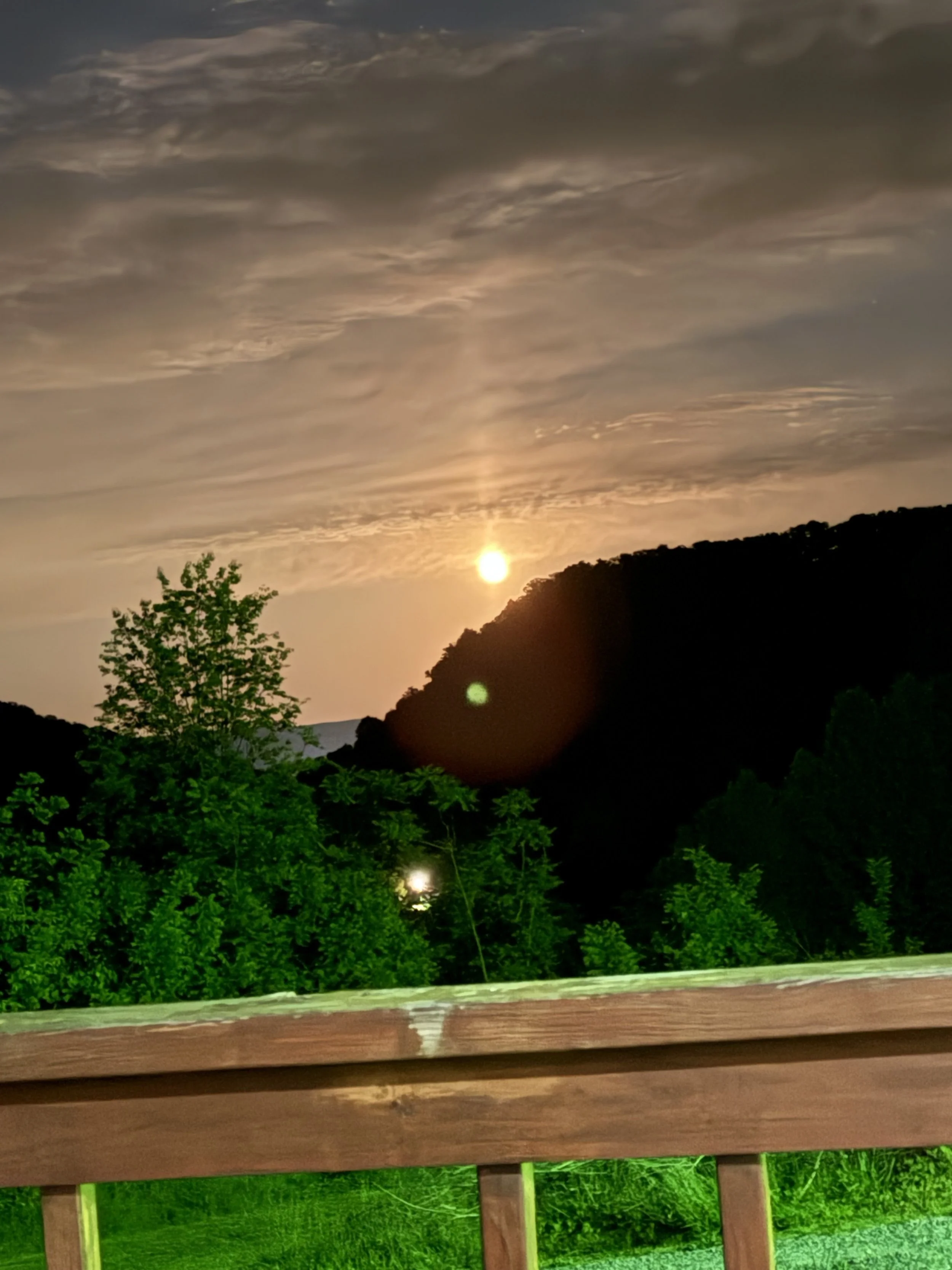 Sunset over a hilly landscape with clouds in the sky and trees in the foreground, viewed from a wooden railing.