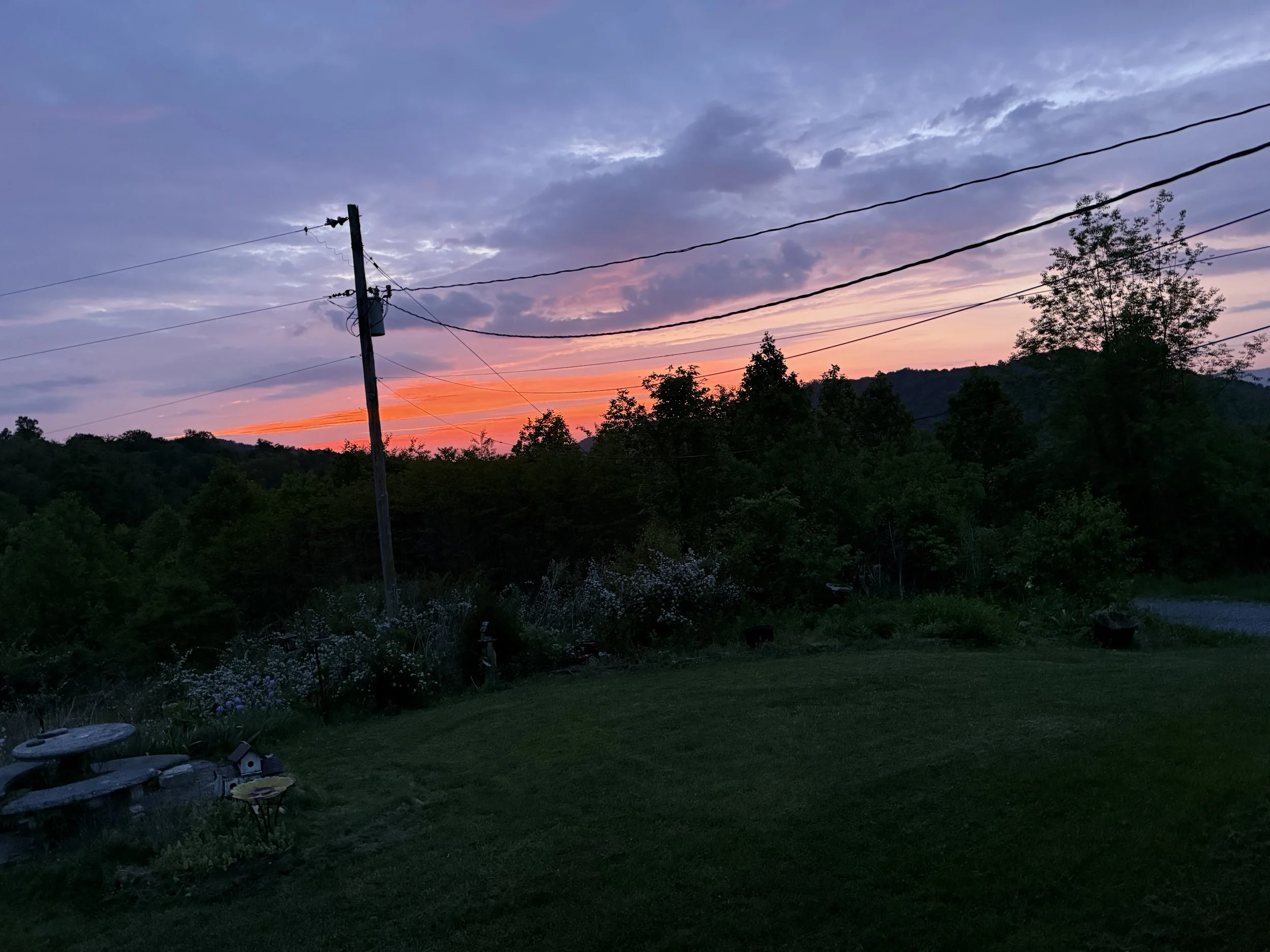 A sunset sky with purple, pink, and orange hues over a rural landscape with trees, power lines, and a utility pole.