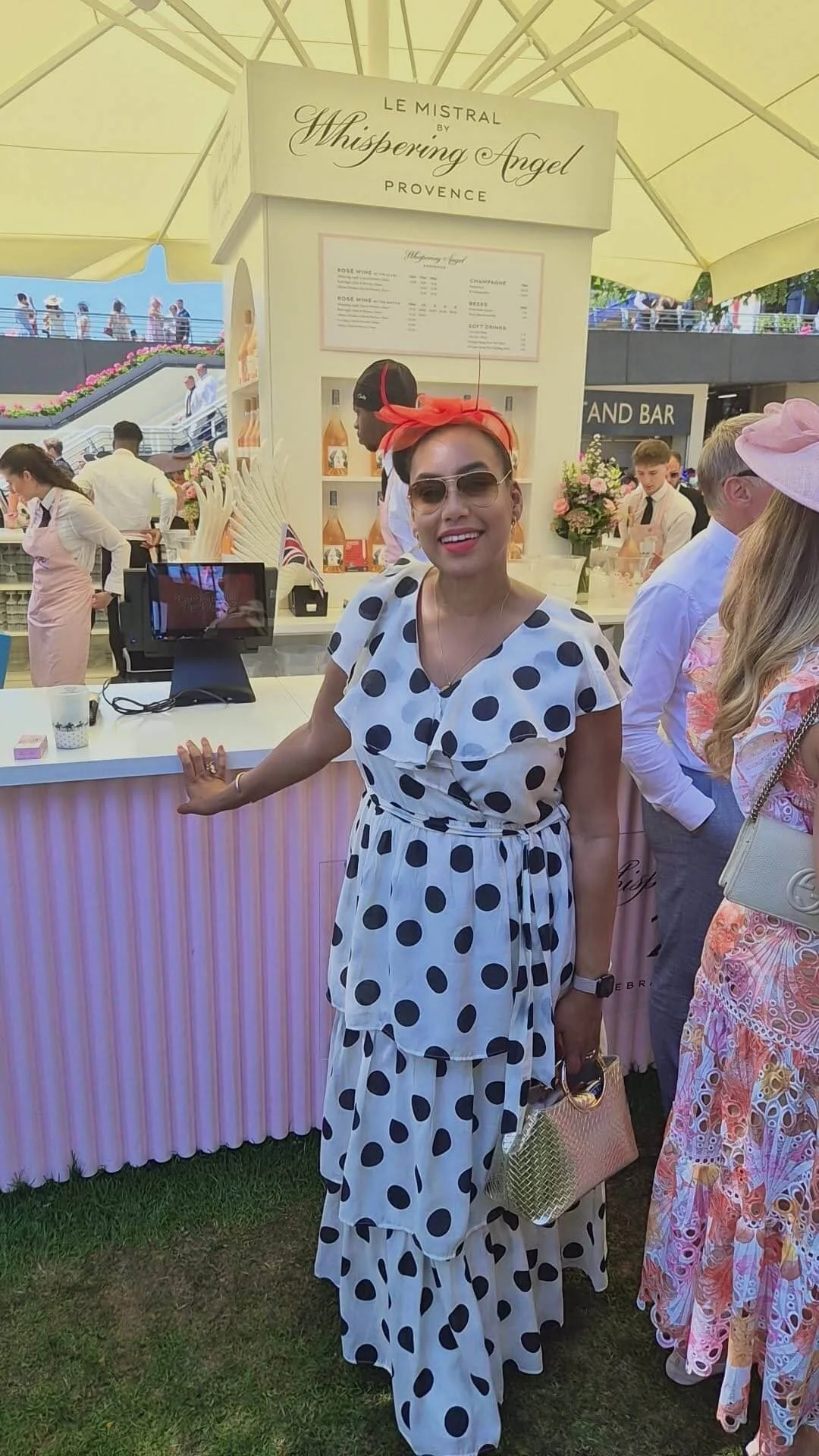 A woman dressed in a polka dot dress and wearing sunglasses, with a red headscarf, standing in front of a pink-striped counter at an outdoor event. There are other people and a lemonade stand in the background.