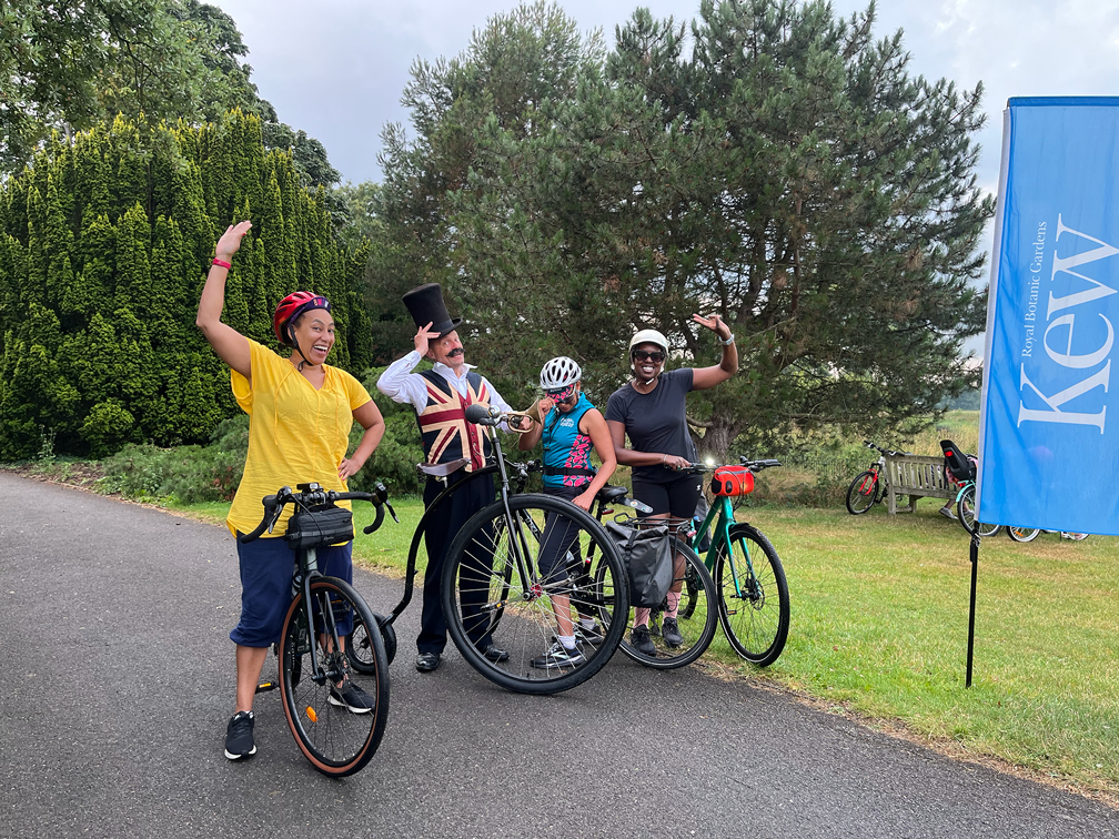 Four people in cycling gear celebrating outdoors, with a person dressed as a circus ringmaster and another wearing a costume with a top hat, standing next to their bikes on a path by a grassy area with trees and a blue sign that reads 'Kew.'