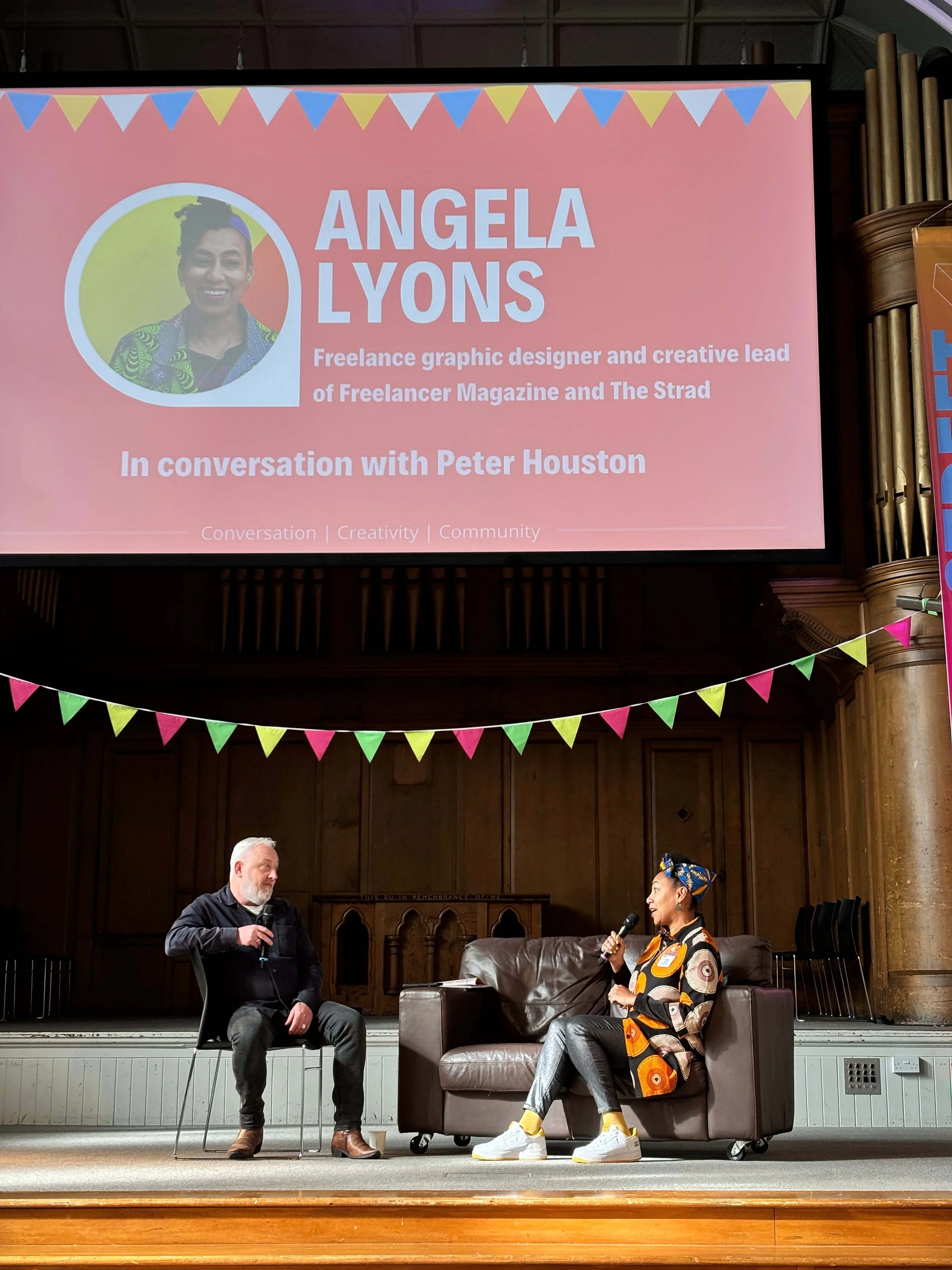 A man and a woman sit on stage engaged in a conversation during an event. The man is holding a microphone, wearing dark clothing, with gray hair and a beard. The woman is holding a microphone, wearing a colorful patterned blazer and yellow sneakers. Behind them, there is a large pink screen displaying the name Angela Lyons, who is a freelance graphic designer and creative lead of Freelancer Magazine and The Strad, in conversation with Peter Houston. The stage is decorated with pink, yellow, and green bunting.