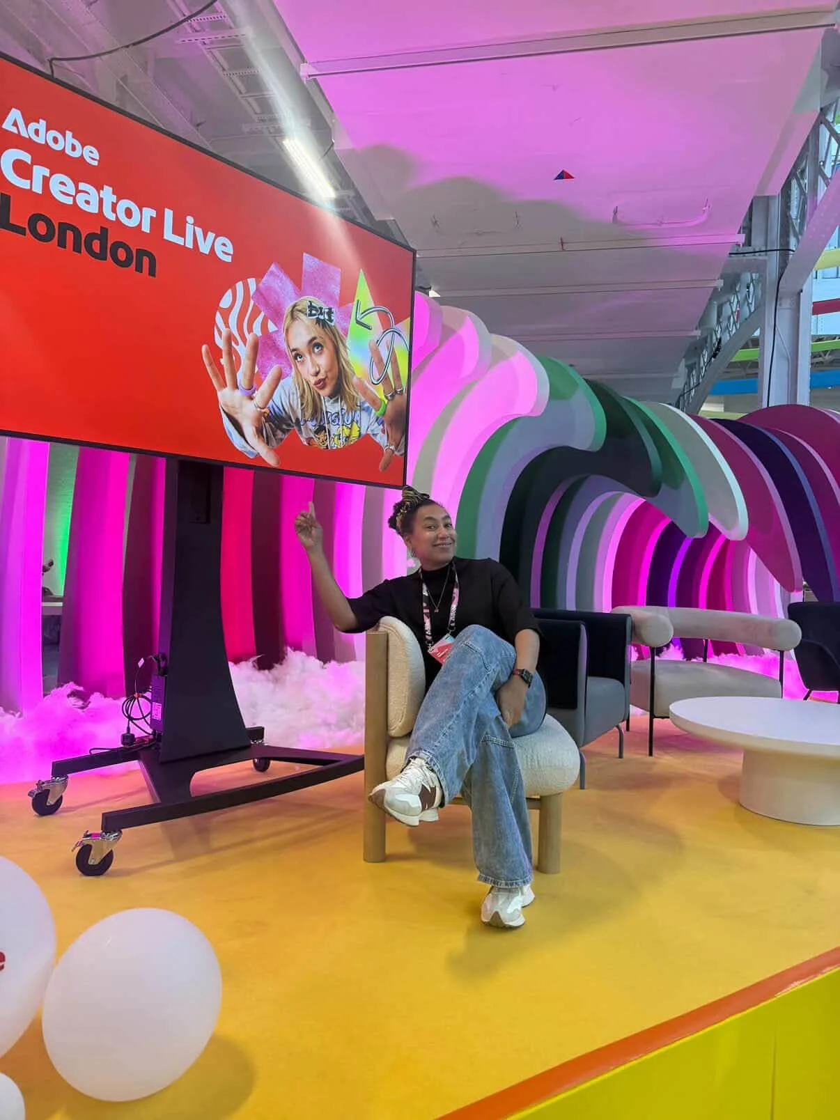 At Adobe Creator Live event in London, a woman sitting in a beige chair, smiling and pointing up, is surrounded by vibrant, colorful background with pink and green wavy structures, pink lighting, and white cloud-like flooring. A large red screen behind her displays the event name and a photo of a young girl with a playful expression.