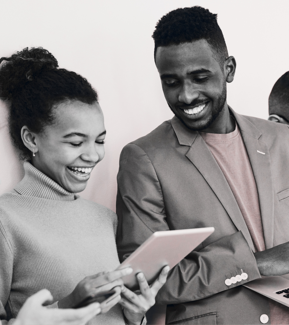 Two people smiling while looking at a tablet, with one holding a laptop. They appear to be in a professional or collaborative setting.