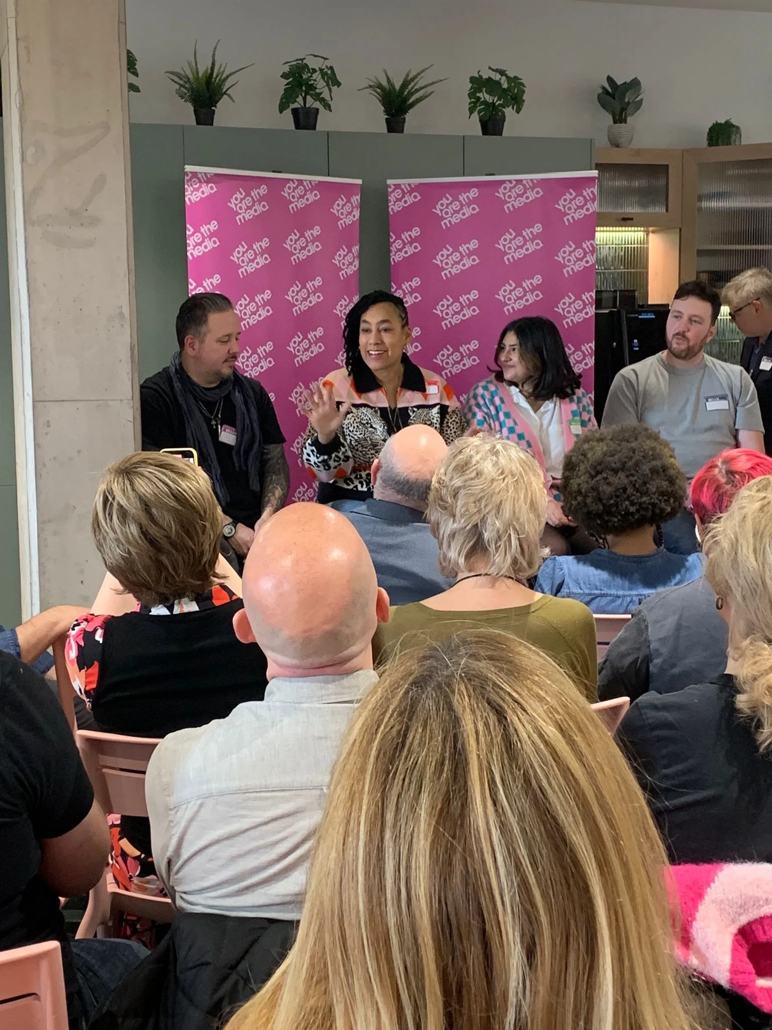 Panel discussion at an event with four women seated at the front and an audience in the foreground. The backdrop features pink banners with the text "You are the media" and potted plants on a shelf behind.