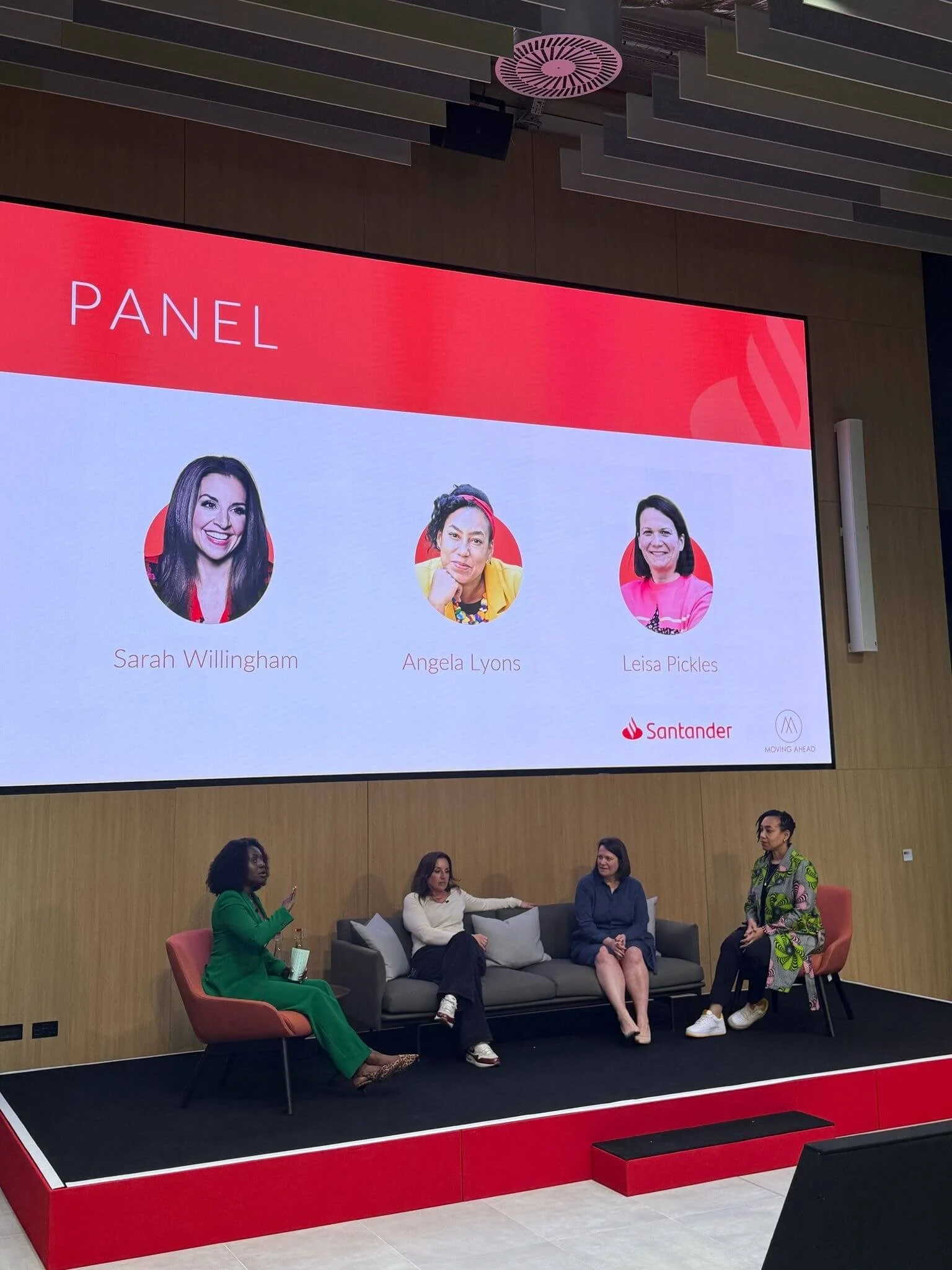 Four women sitting and standing on a stage with a large screen behind them. The screen displays images of three women and the word 'PANEL' at the top, with logos of Santander and Moving Ahead at the bottom.