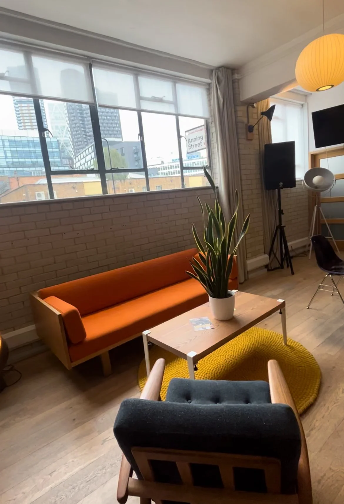 Modern living room with an orange sofa, wooden coffee table with a potted plant, retro armchair, and large windows with city view.