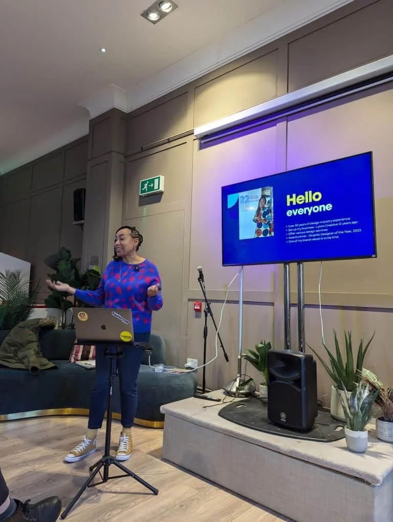 Woman giving a presentation in a room with a large screen displaying a slide that says "Hello everyone" and a colorful image. She is standing near a laptop on a stand, gesturing with her right hand, with a microphone nearby. Potted plants are on a table next to her, and a couch with some belongings behind her.