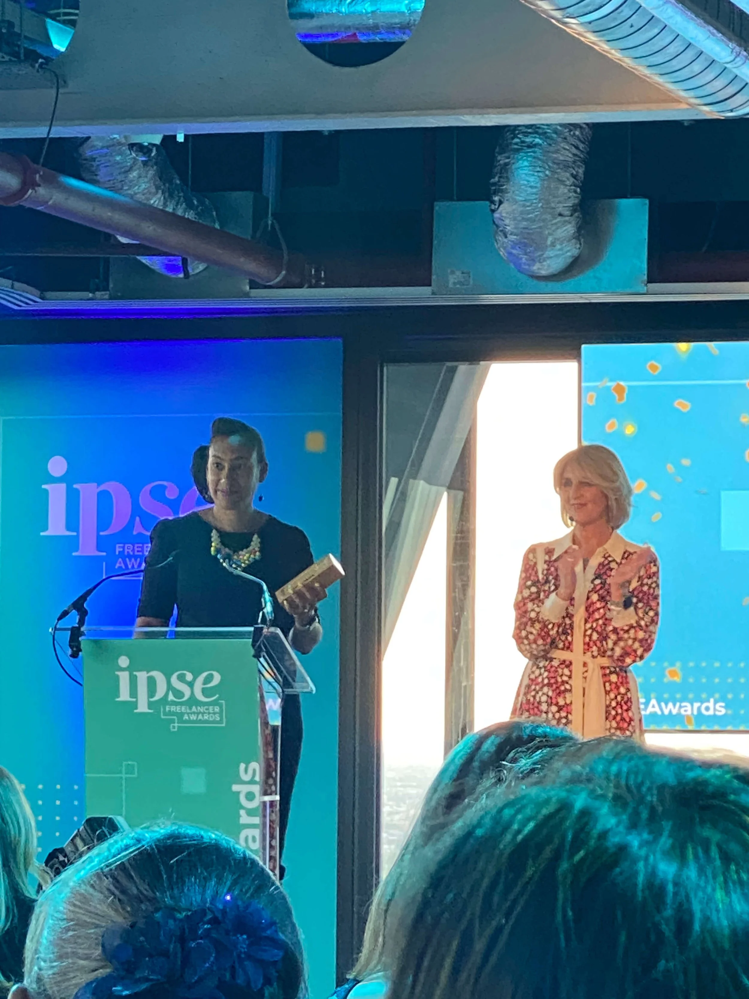 Two women standing on stage at the IPSE Freelance Awards, with one woman speaking at a podium and the other holding a trophy. There are photographers and audience members in front, with balloons and a blue backdrop.