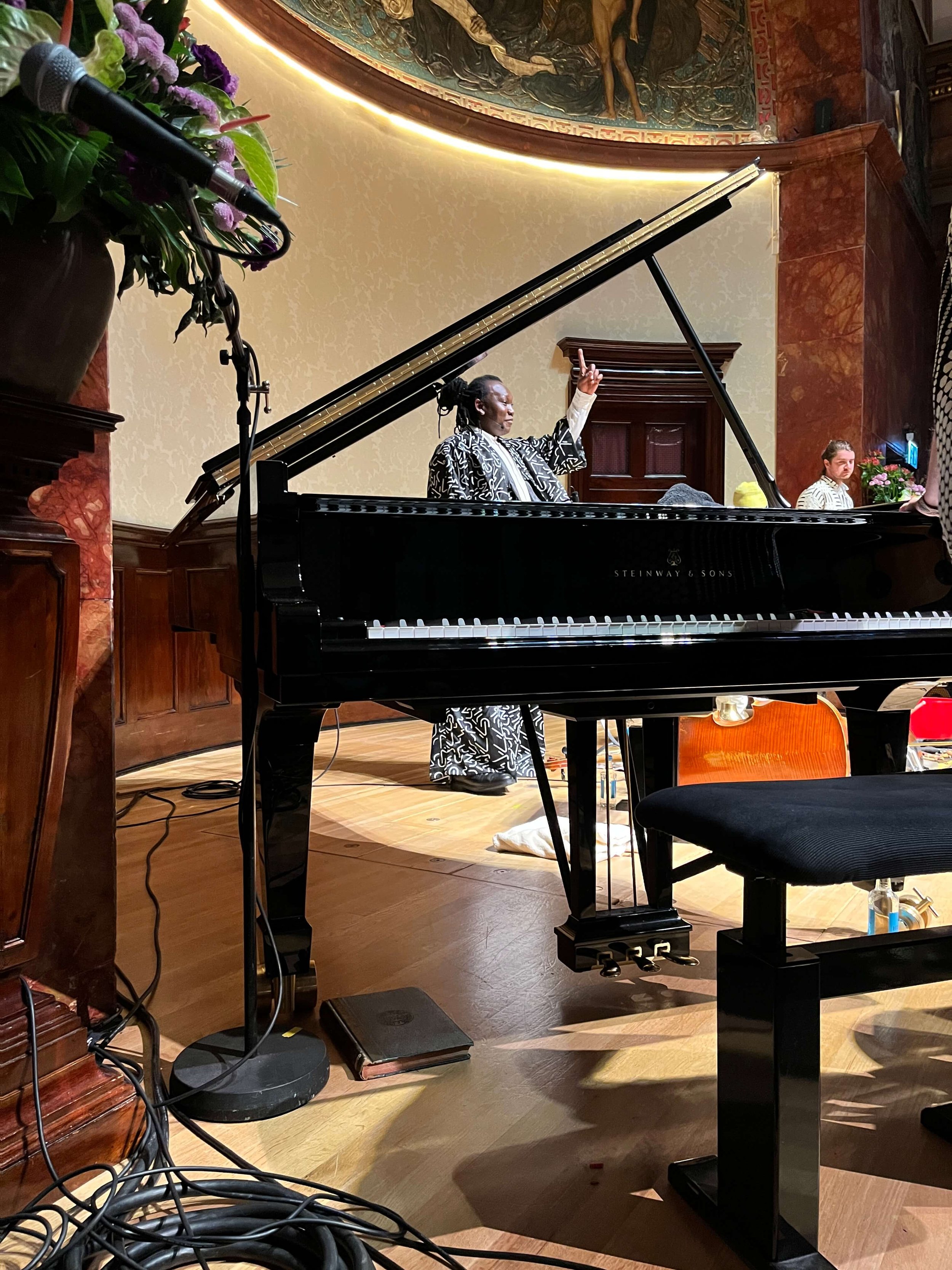 Abel Selaocoe performing with a piano, raising one finger, in a venue decorated with flowers, an ornate ceiling, and wooden wall paneling.