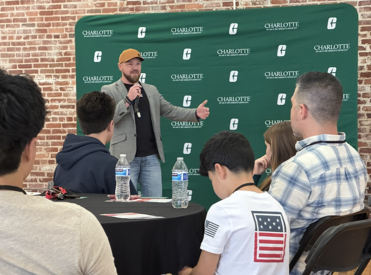 A man with a beard, gray blazer, black shirt, and yellow cap speaking into a microphone in front of a green backdrop with 'Charlotte' and a logo; an audience of mixed teens and adults listening, two water bottles on the table.