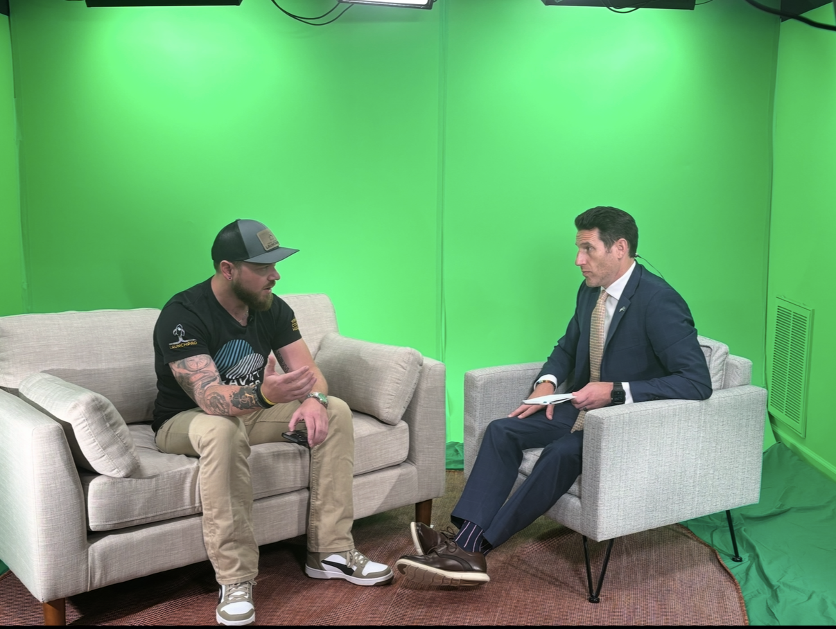A man in casual clothing with tattoos, wearing sneakers and a baseball cap, speaks during an interview with a man in a dark suit and tie who is holding notes, set in a studio with a green screen background.