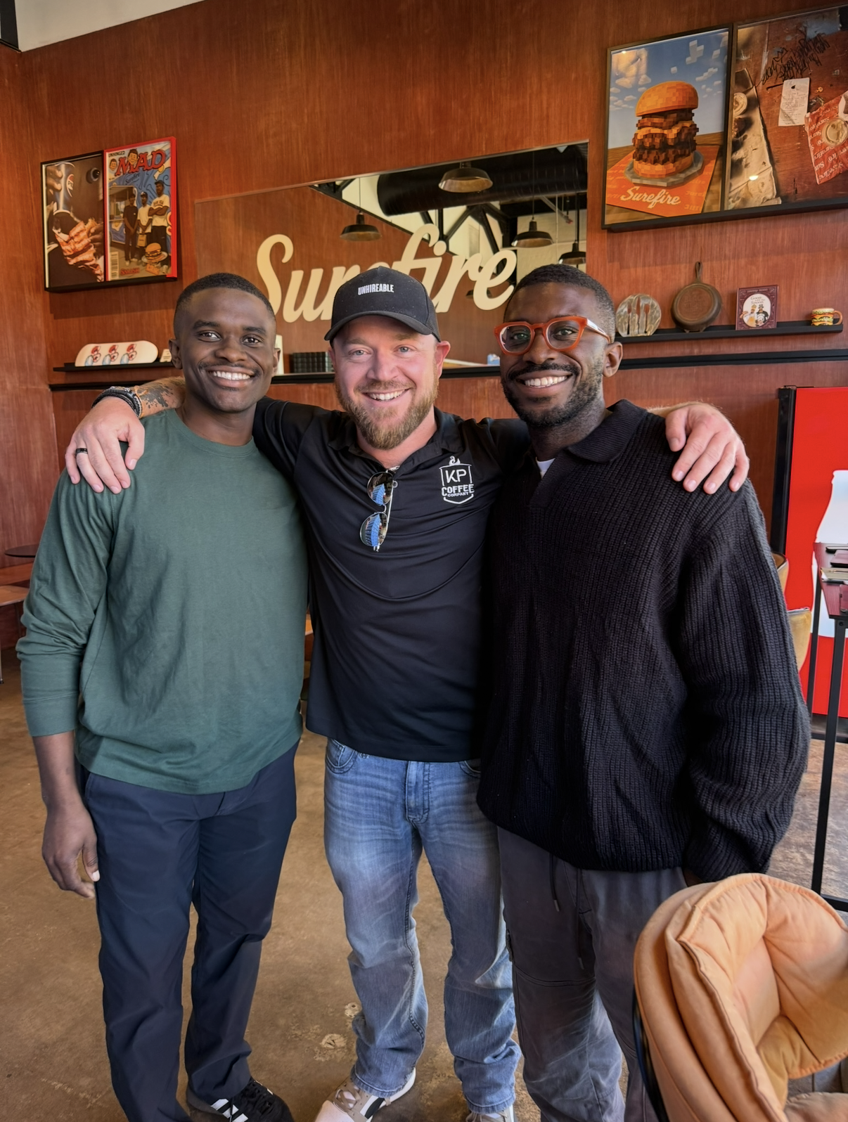 Three men smiling and standing close together in a coffee shop, with arms around each other. The background features wood paneling, framed pictures, and the word 'Surefire' on the wall.
