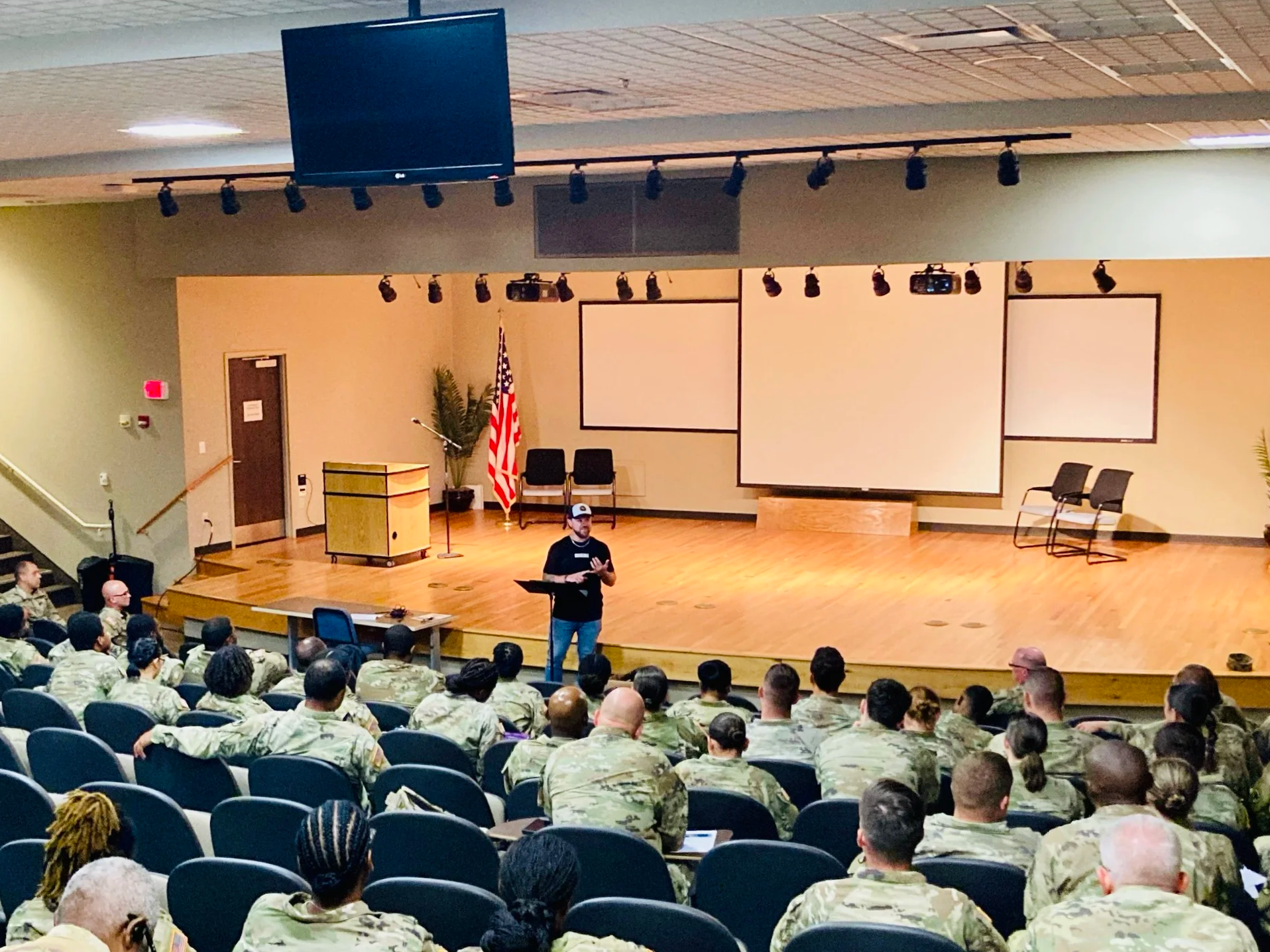 A speaker stands on a stage in front of a military audience inside an auditorium, with American flags and presentation screens behind him.