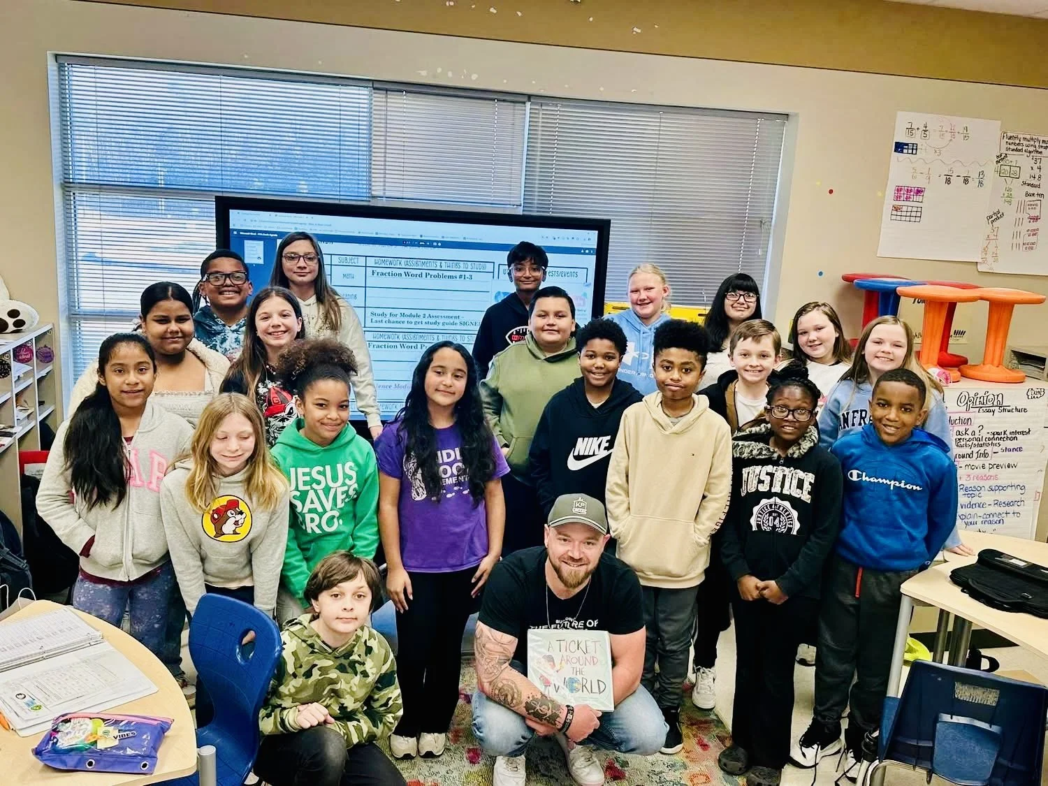 A classroom with students and a teacher, all smiling, standing in front of a digital display. The teacher is kneeling in front, holding a book titled "A Ticket Around the World." The classroom has desks, colorful chairs, and educational posters on the walls.