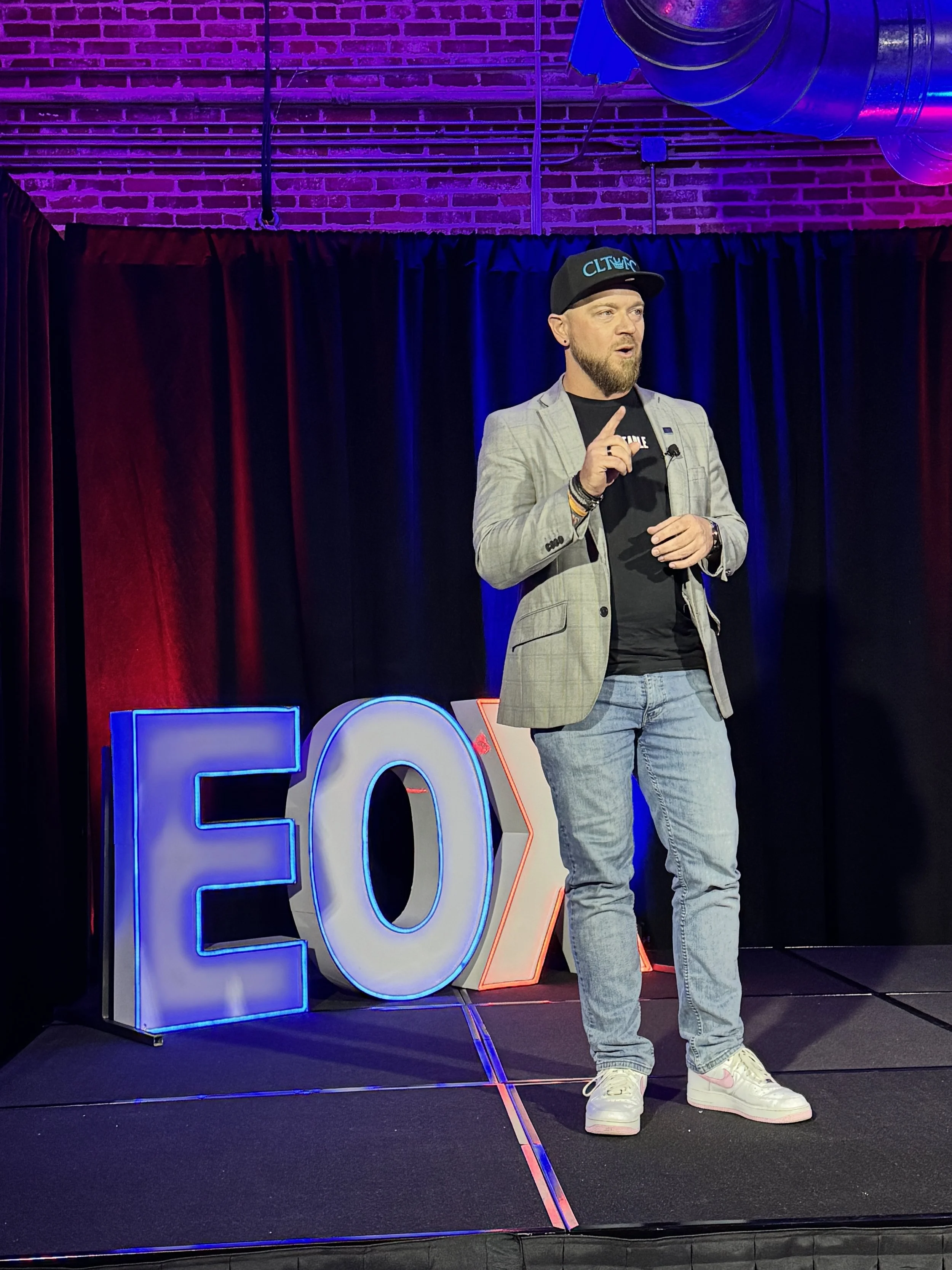 A man with a beard and short hair wearing a black baseball cap, a gray blazer, a black t-shirt, ripped jeans, and white sneakers is standing on stage in front of a black curtain with purple, blue, and red lighting. He is gesturing with his right index finger and holds a microphone near his chest. To his left, there are large illuminated numbers '2' and '0' in red and blue respectively.