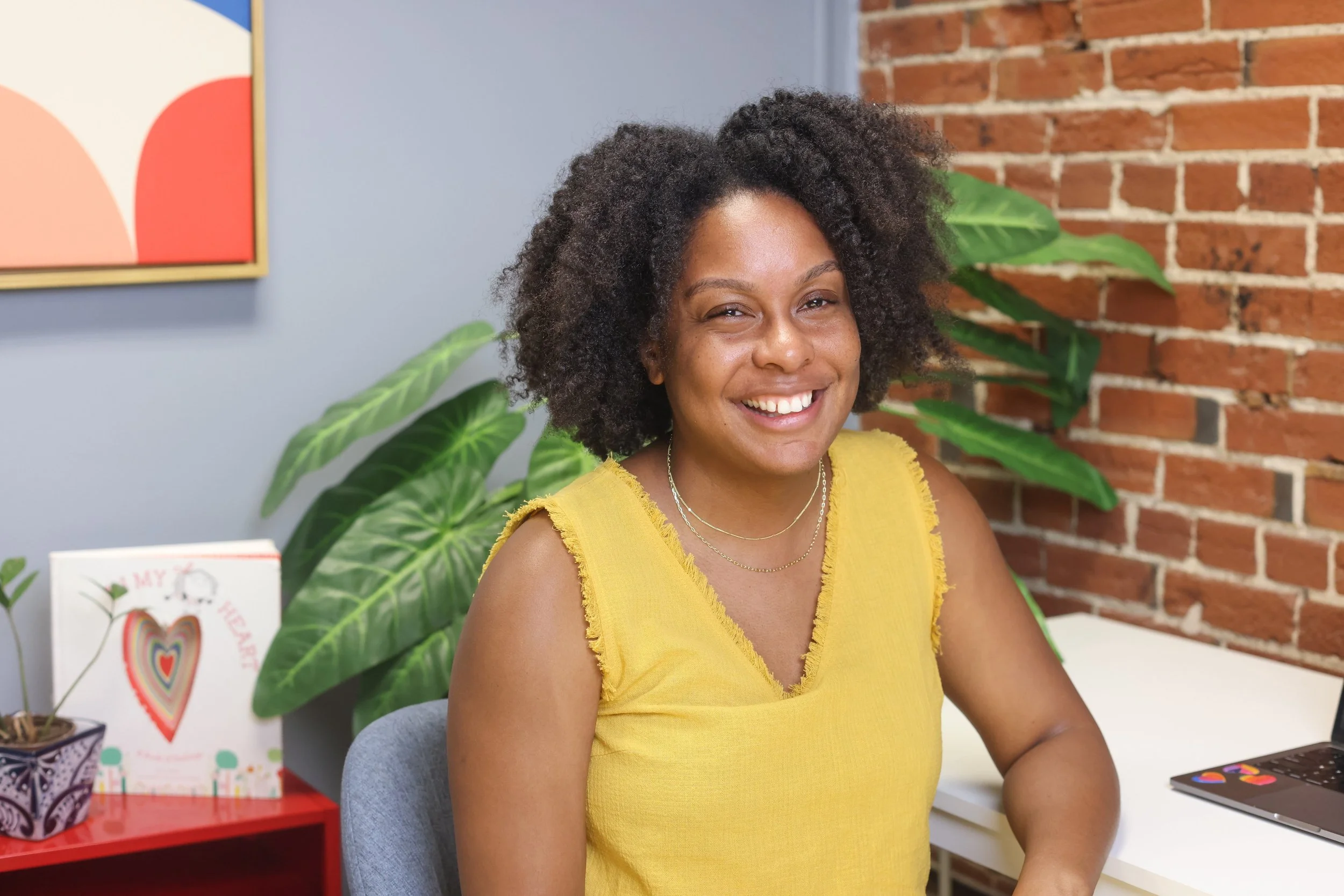 A Black therapist wears a yellow tank top, short afro, and a big smile while sitting in her office. Learn play therapy techniques from one of the Midwest's growing leaders in child therapy.