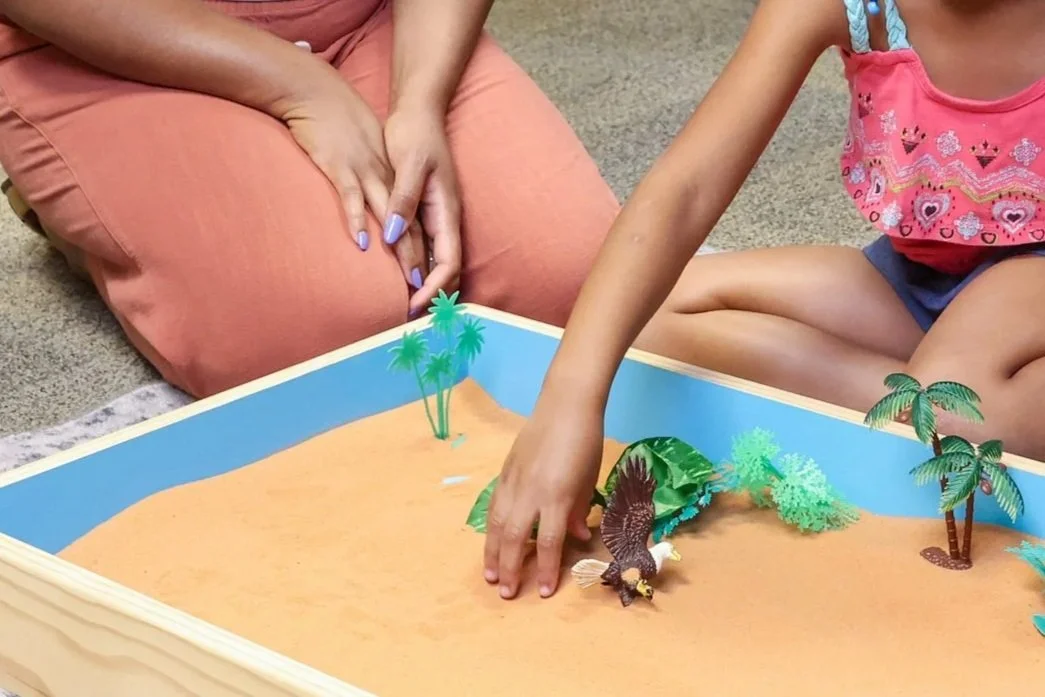 A Black child's hand arranges miniature figures in a sand tray box. Play therapy can be a fun and supportive way for your child to process big feelings, learn coping skills, and become confident. Get started in St. Louis today!
