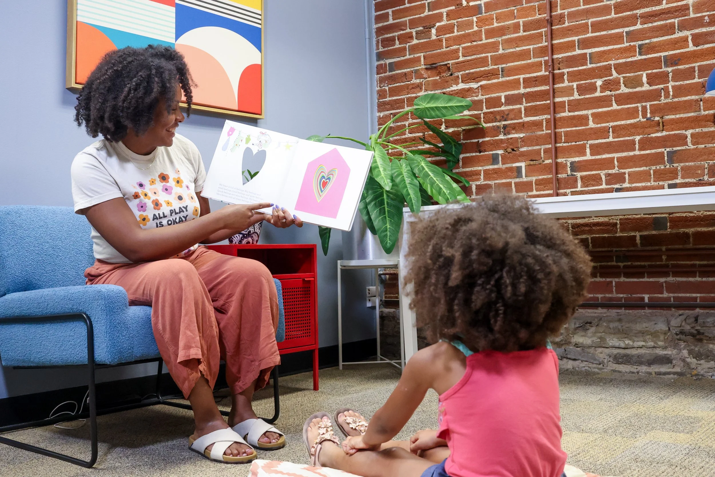 A Black woman therapist holds a book and smiles at a Black child sitting on the floor. Children can have fun while learning emotional regulation skills in play therapy in St. Louis.