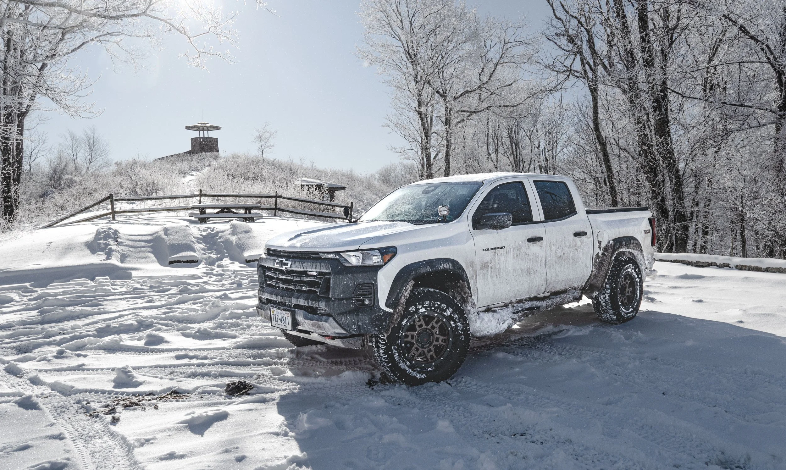 White pickup truck parked on a snow-covered landscape surrounded by snowy trees, with hills and a structure in the background.