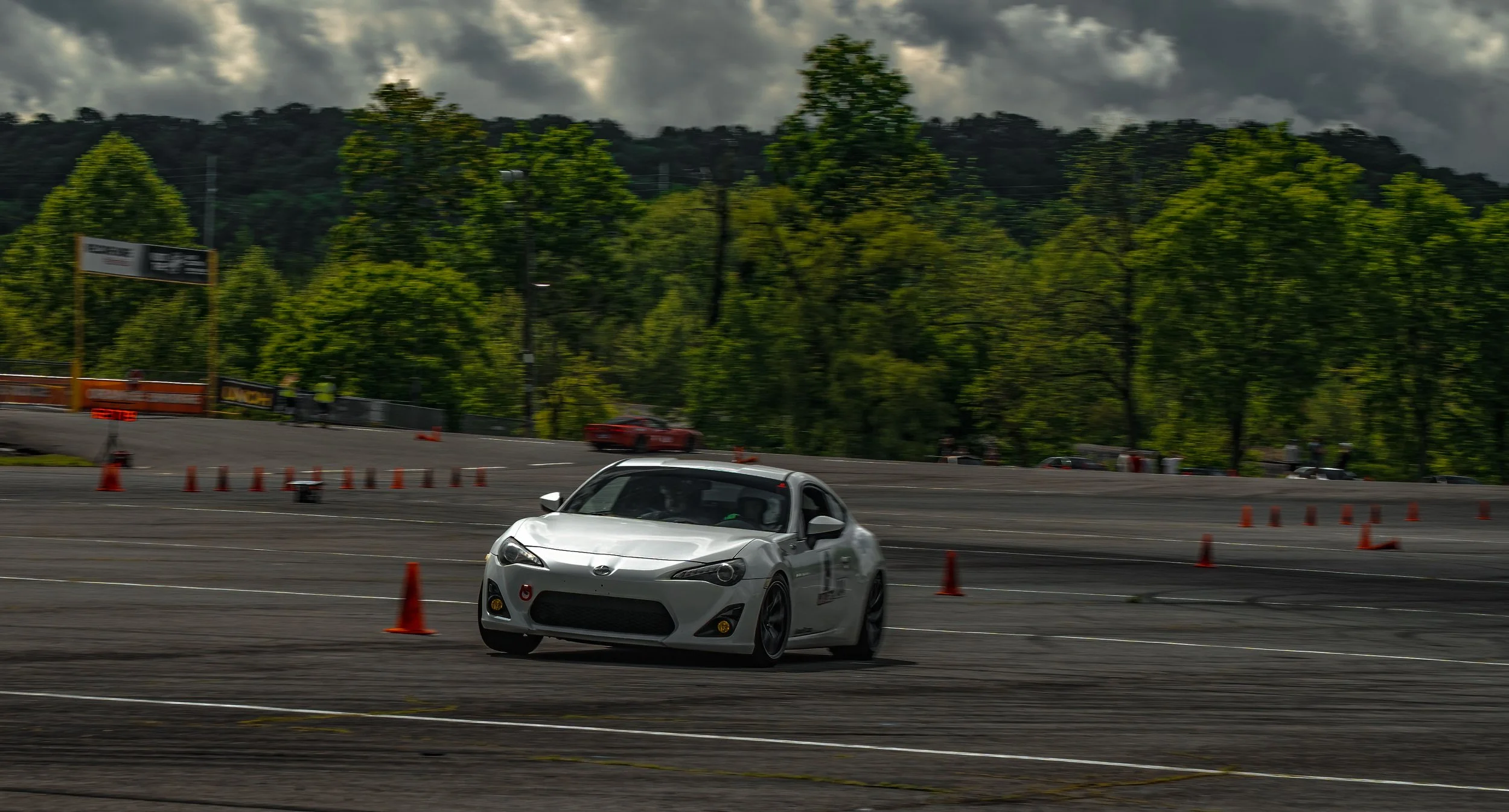 A white sports car navigating through a slalom course marked by orange traffic cones on a paved lot, with trees and hills in the background under cloudy skies.