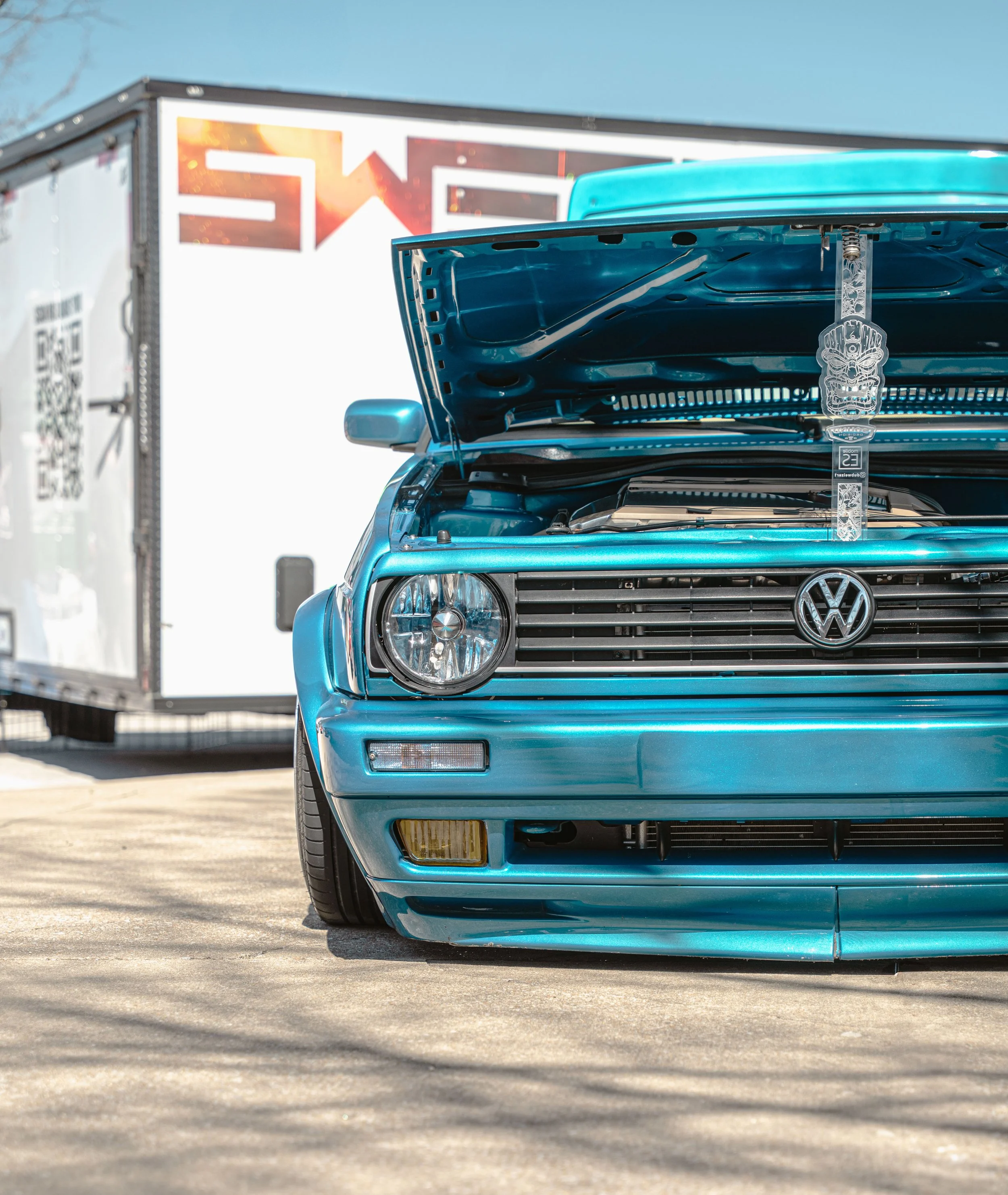 Blue Volkswagen car with open hood parked on concrete with a white trailer in the background.