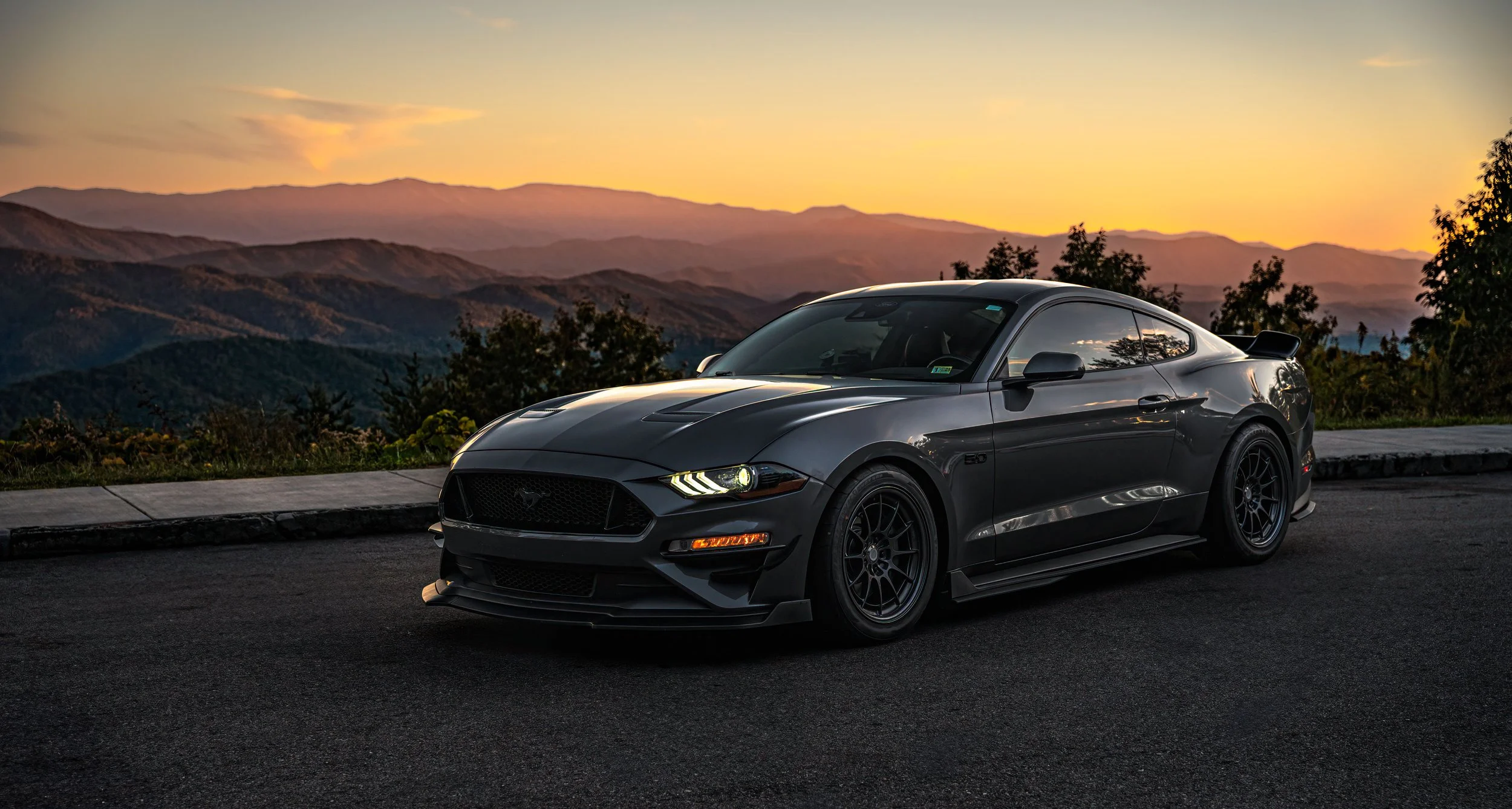 A gray Ford Mustang GT car parked on a mountain road at sunset with a backdrop of mountains and a colorful sky.